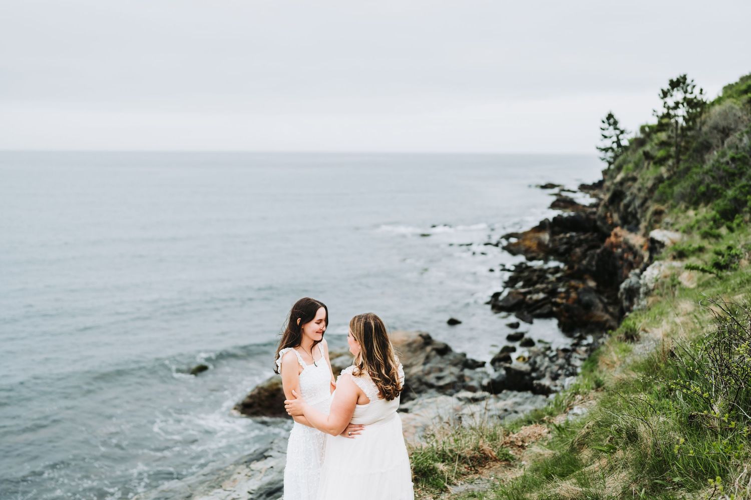 A romantic couple in white embraces on a rocky coastal cliff overlooking the misty ocean.
