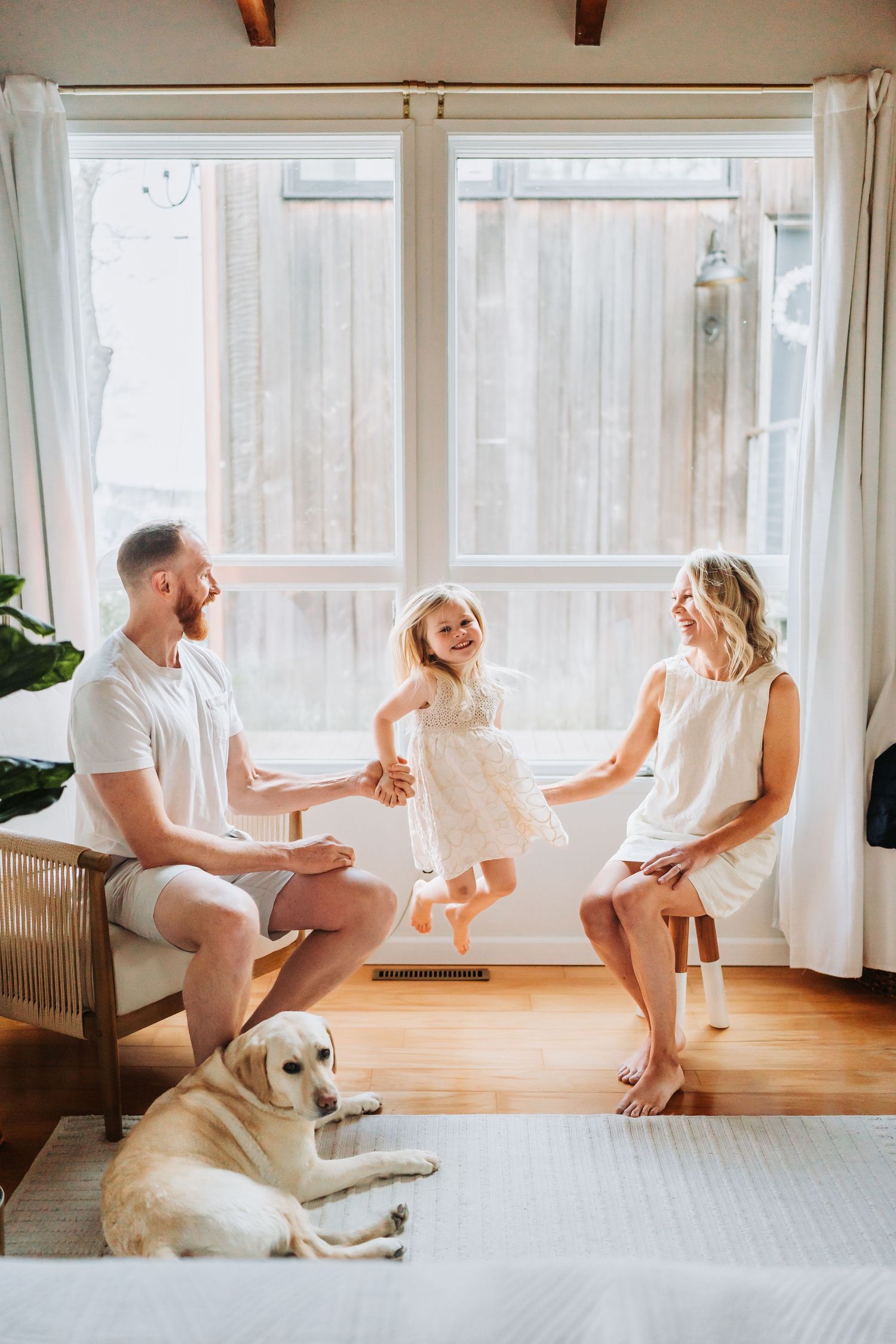 Joyful family with a young girl jumping between parents while a golden Labrador rests nearby.