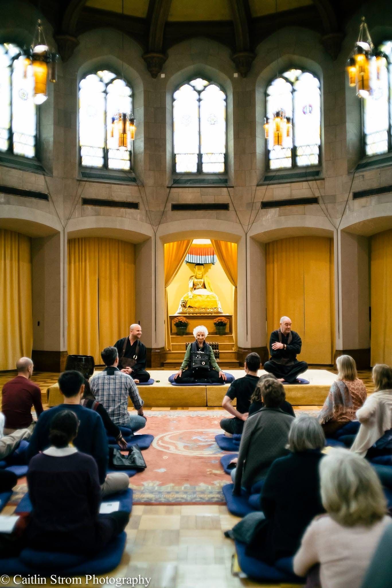 People gathered in a circular room with gothic windows and yellow curtains for a meditation or spiritual session.