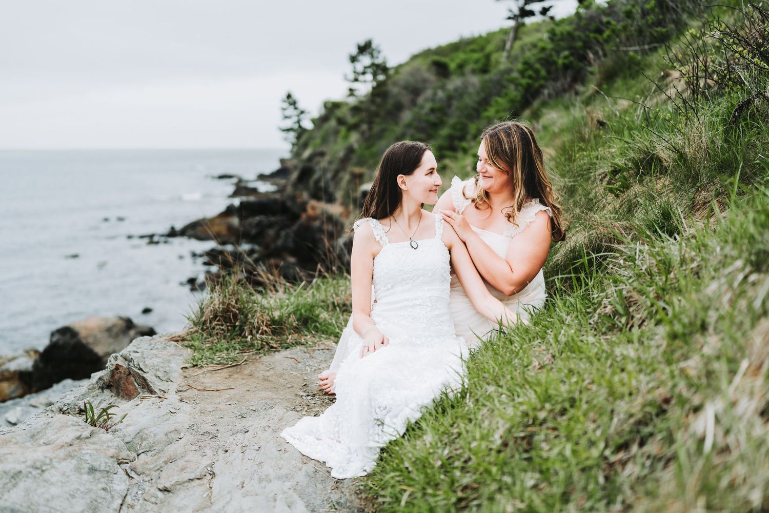 Two people in white dresses embrace on a scenic coastal cliff overlooking the ocean on a cloudy day.