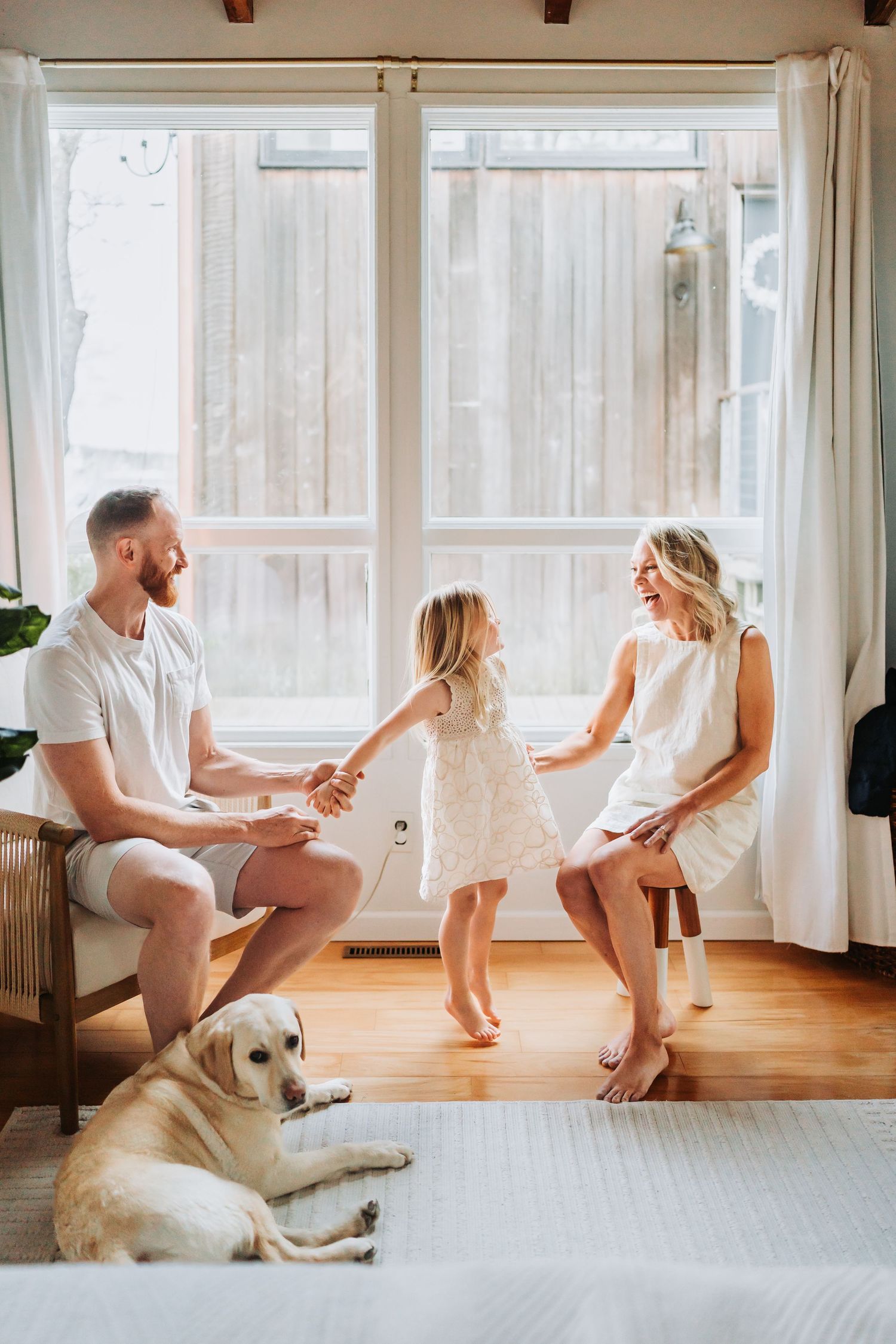 A happy family with a young girl dancing between parents and a golden Labrador resting on hardwood floors near large windows.