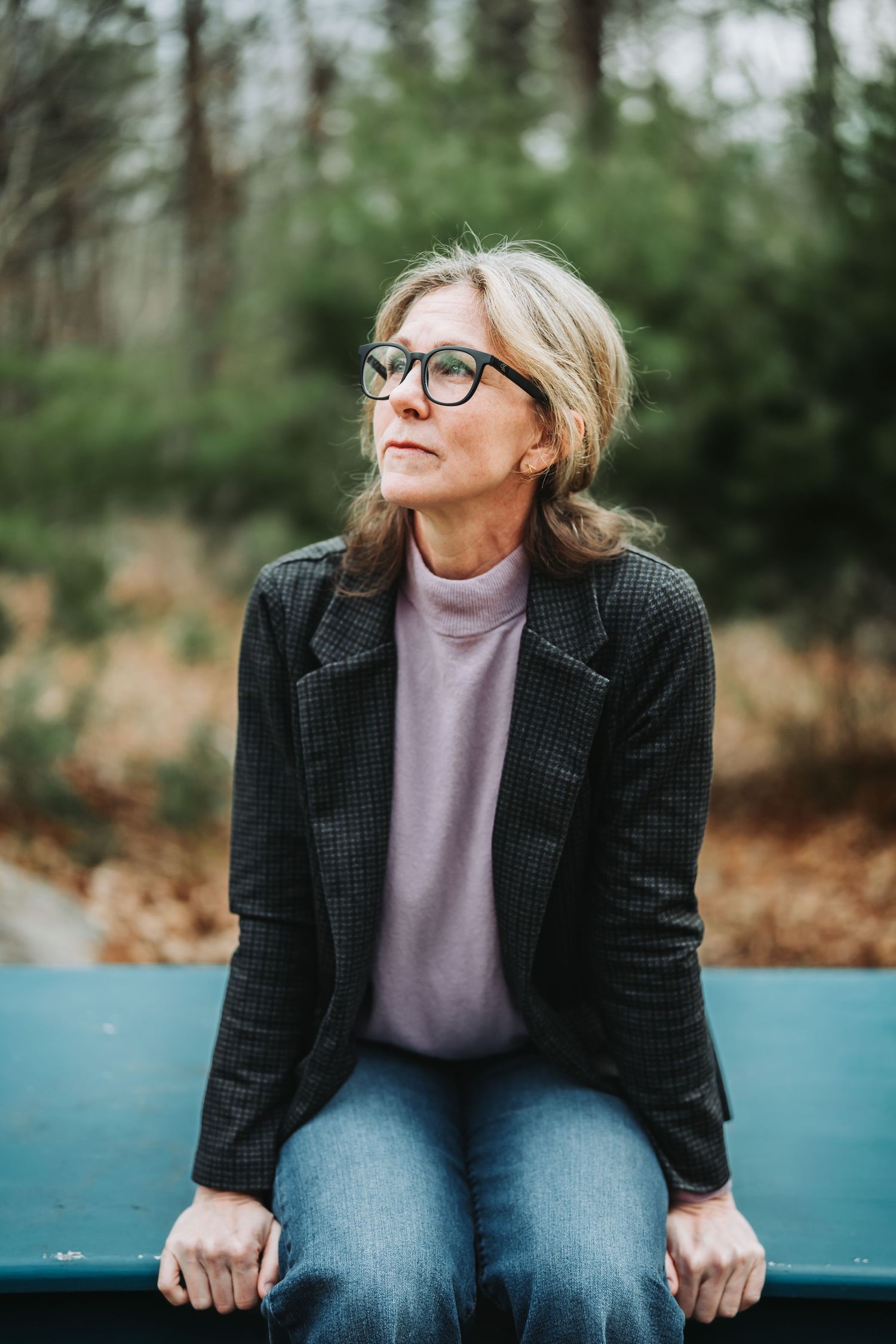 Person in black blazer and lavender top sits thoughtfully on blue bench in autumn outdoor setting.