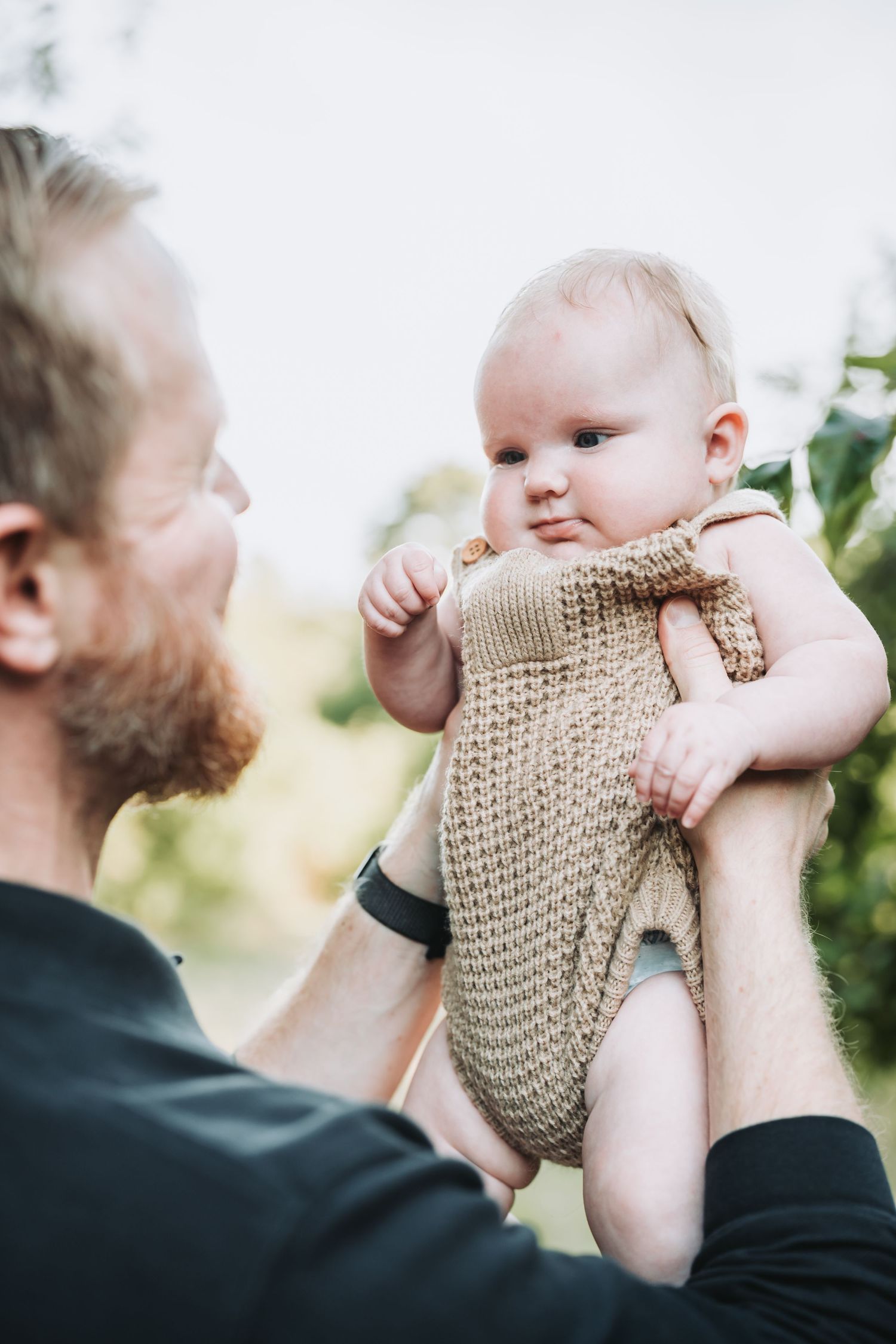 Baby in beige knit outfit being lifted up against bright outdoor background.