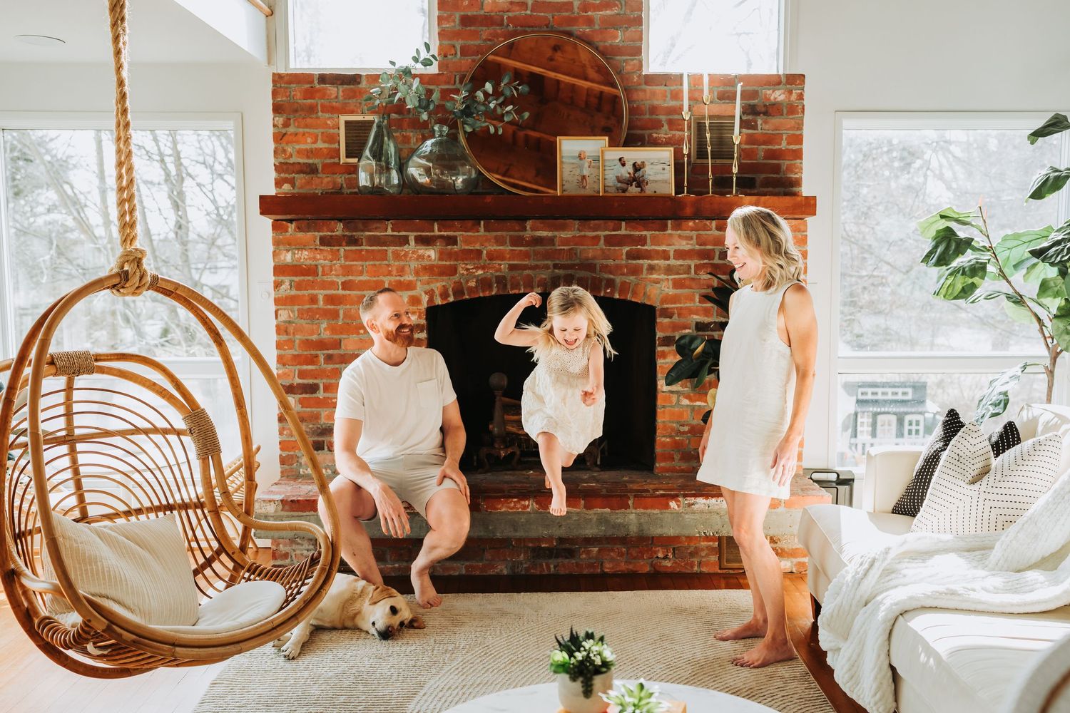Family enjoying a cozy living room with brick fireplace, rattan swing chair, and child jumping with joy.