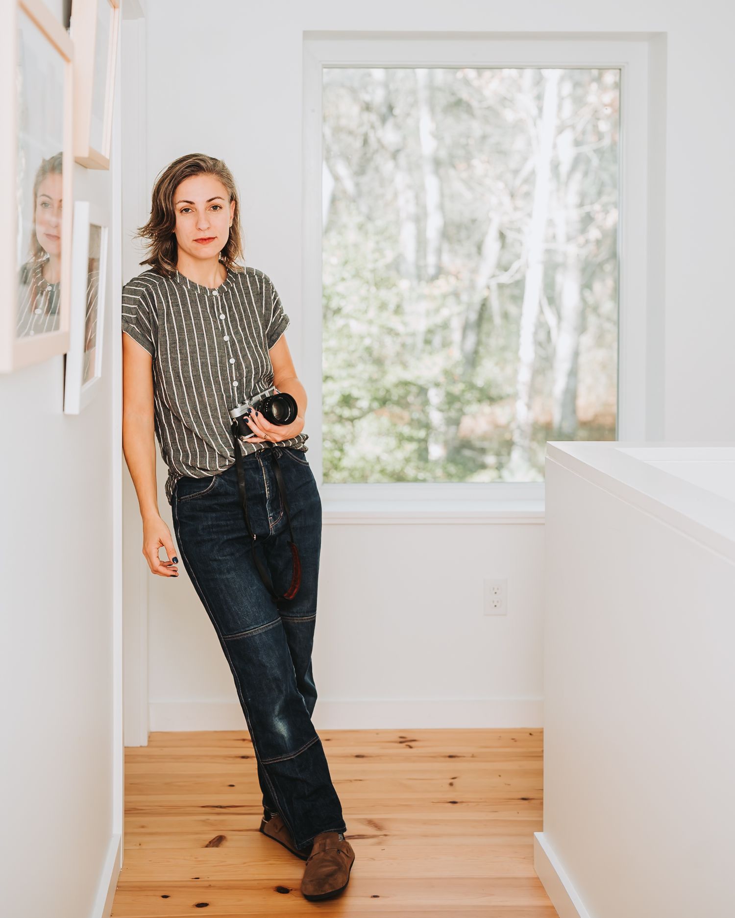 Person in striped top and dark jeans sits casually in a bright, modern room with wooden flooring and white walls.