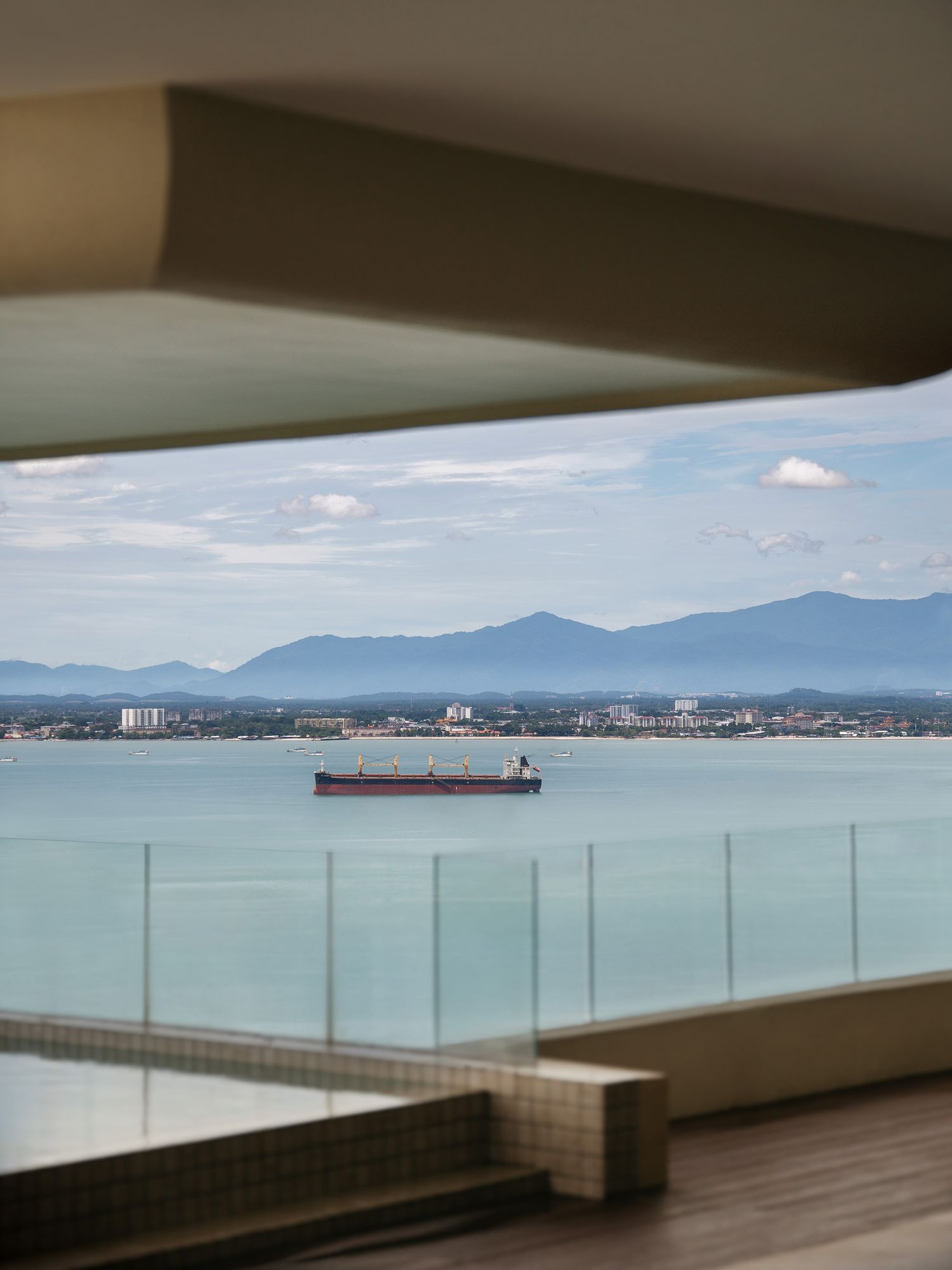 Panoramic ocean view through a modern balcony with glass railings overlooking a coastal city and mountains in the distance.