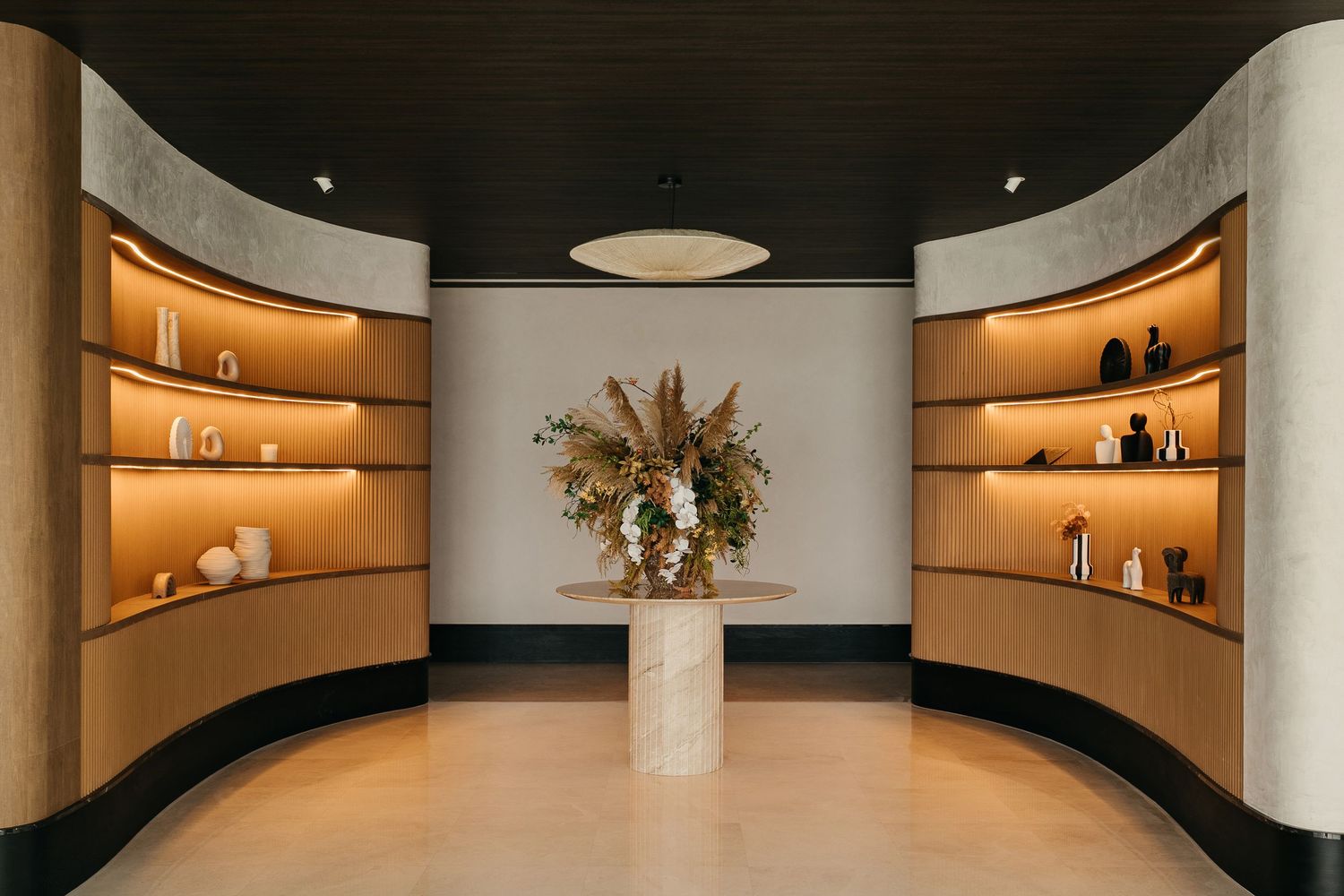 Modern interior space with curved wooden display shelves illuminated by warm lighting and featuring a dried floral arrangement.