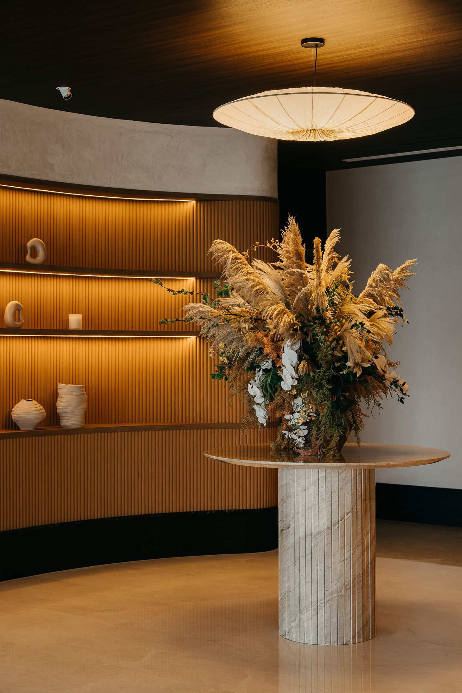 Modern minimalist lobby interior with marble column featuring dried pampas grass arrangement under paper lantern lighting.
