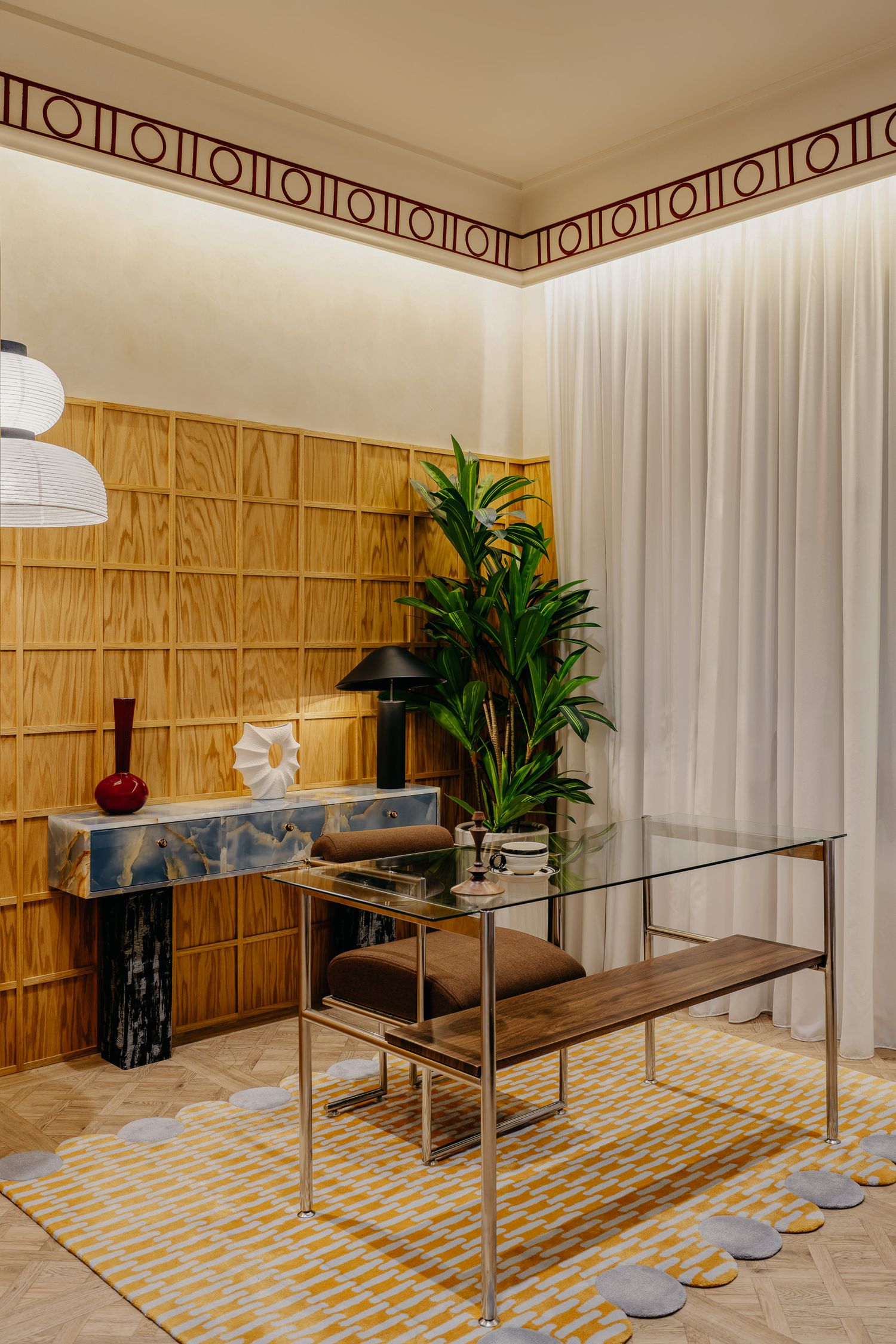 Modern dining space with glass table and brown seating against wood-paneled wall with geometric border trim.