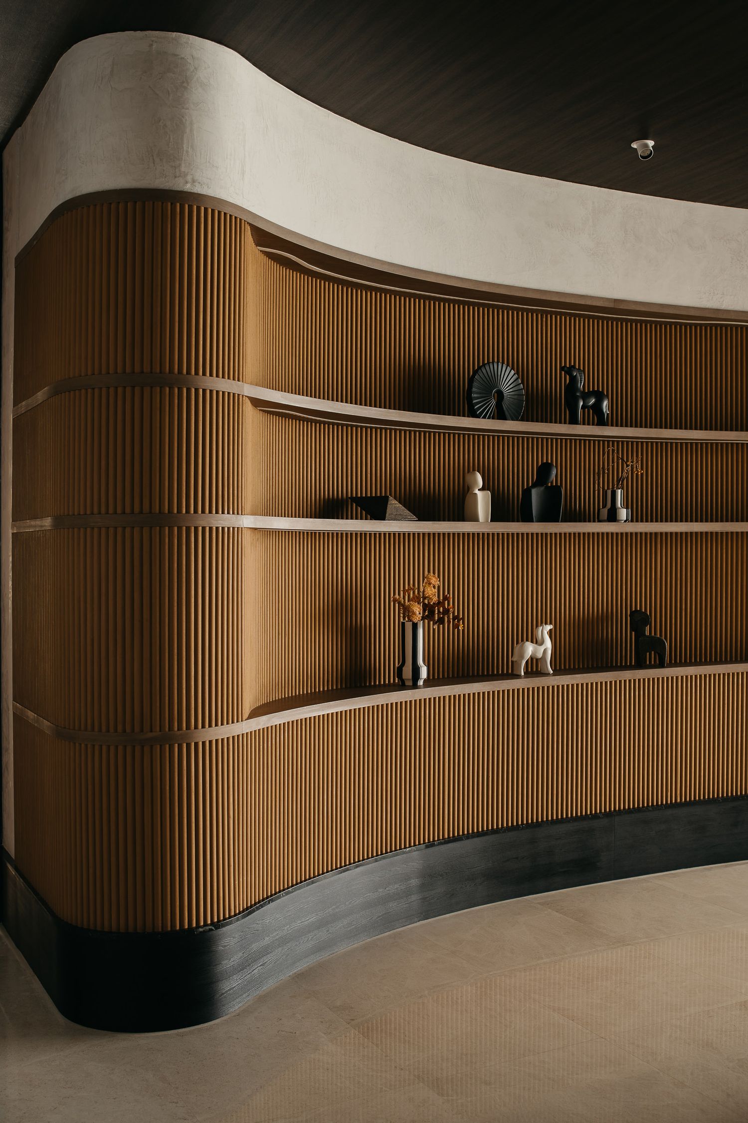 Modern interior design featuring curved wooden slat wall with floating shelves displaying decorative objects in warm lighting.