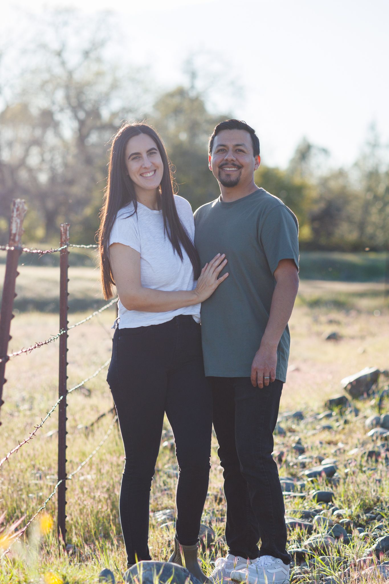 A couple stands together outdoors in casual attire against a natural landscape with barbed wire fence.