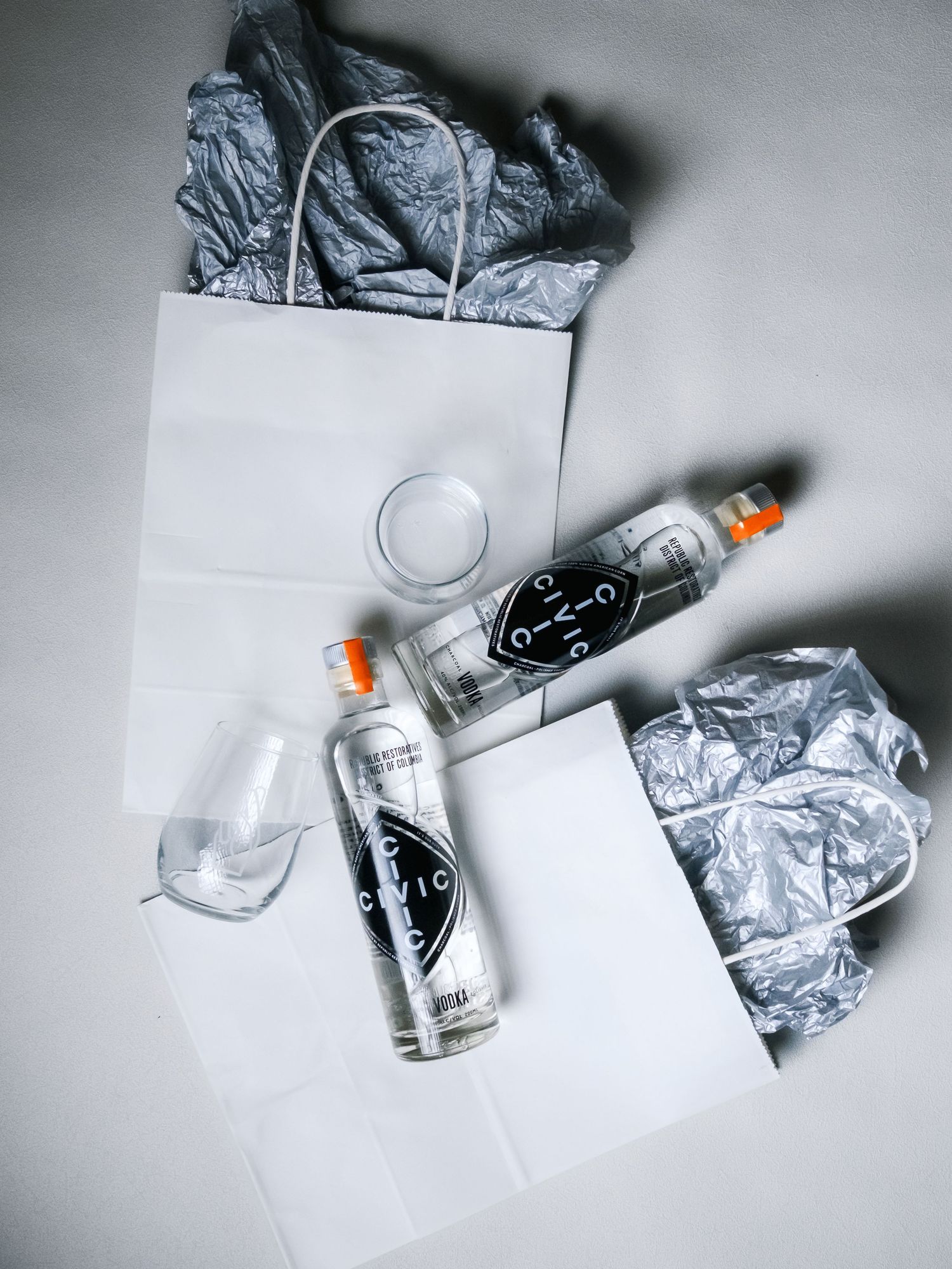 Flatlay arrangement of clear glass bottles and crumpled silver paper with white shopping bags on a light surface.