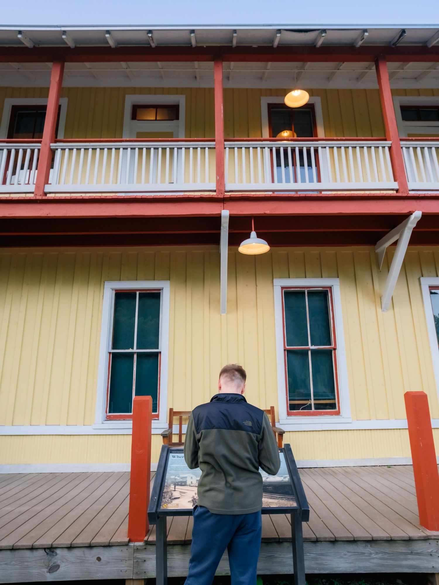 A person views a train station platform through a large window of a yellow building with a covered porch walkway.