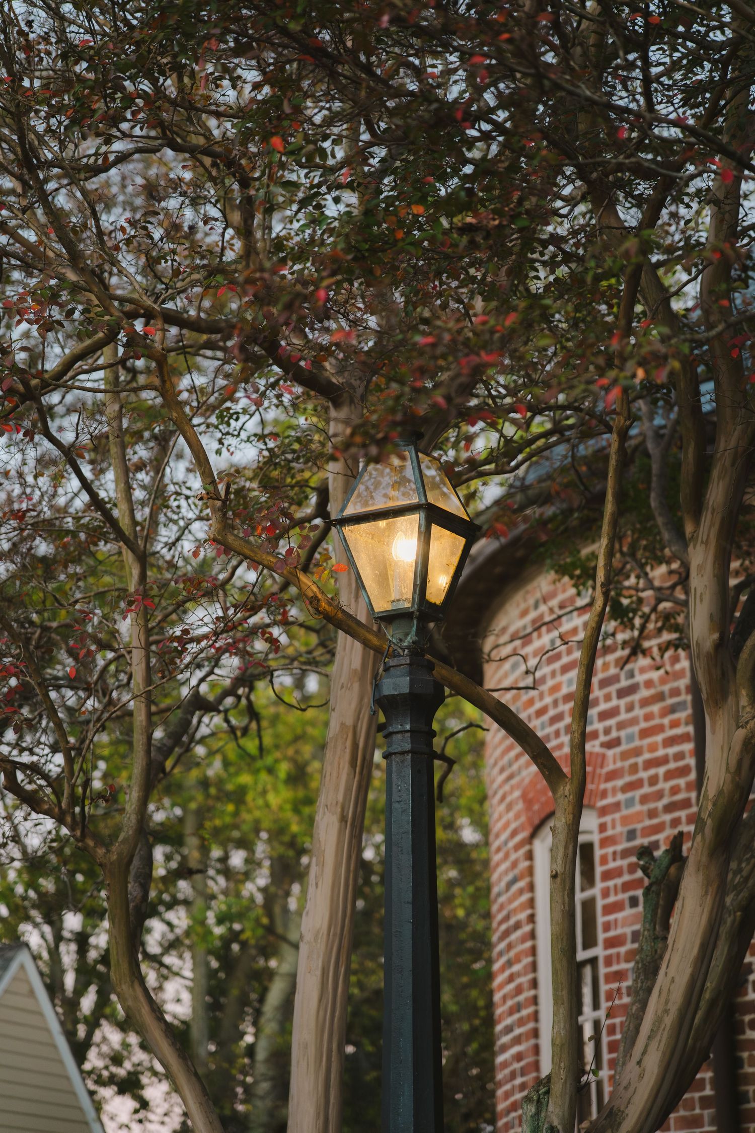 An illuminated vintage street lamp glows against autumn trees and a red brick building with arched windows.