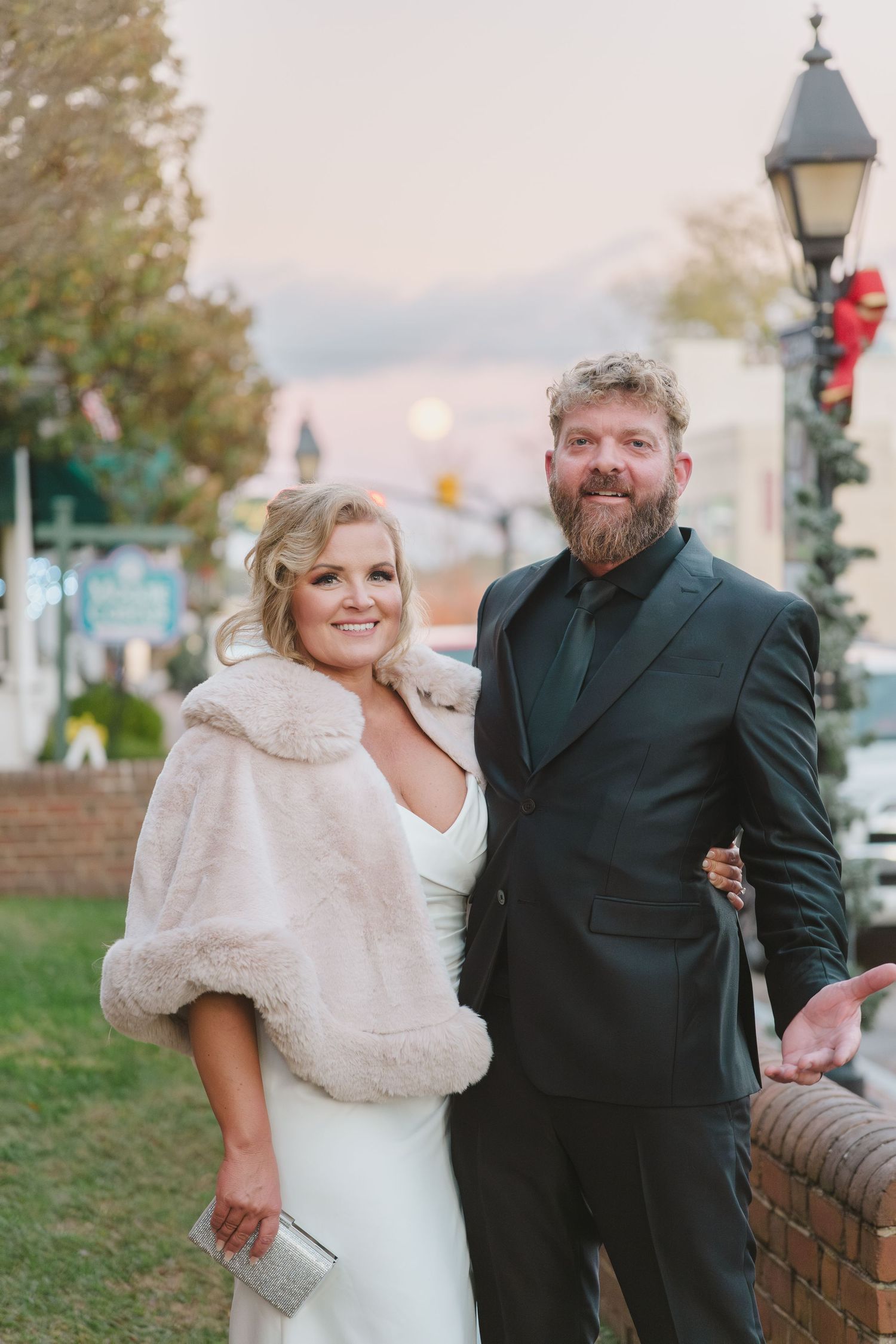 A newlywed couple stands together outdoors near a lamp post at dusk, the bride wearing a white dress and fur wrap.