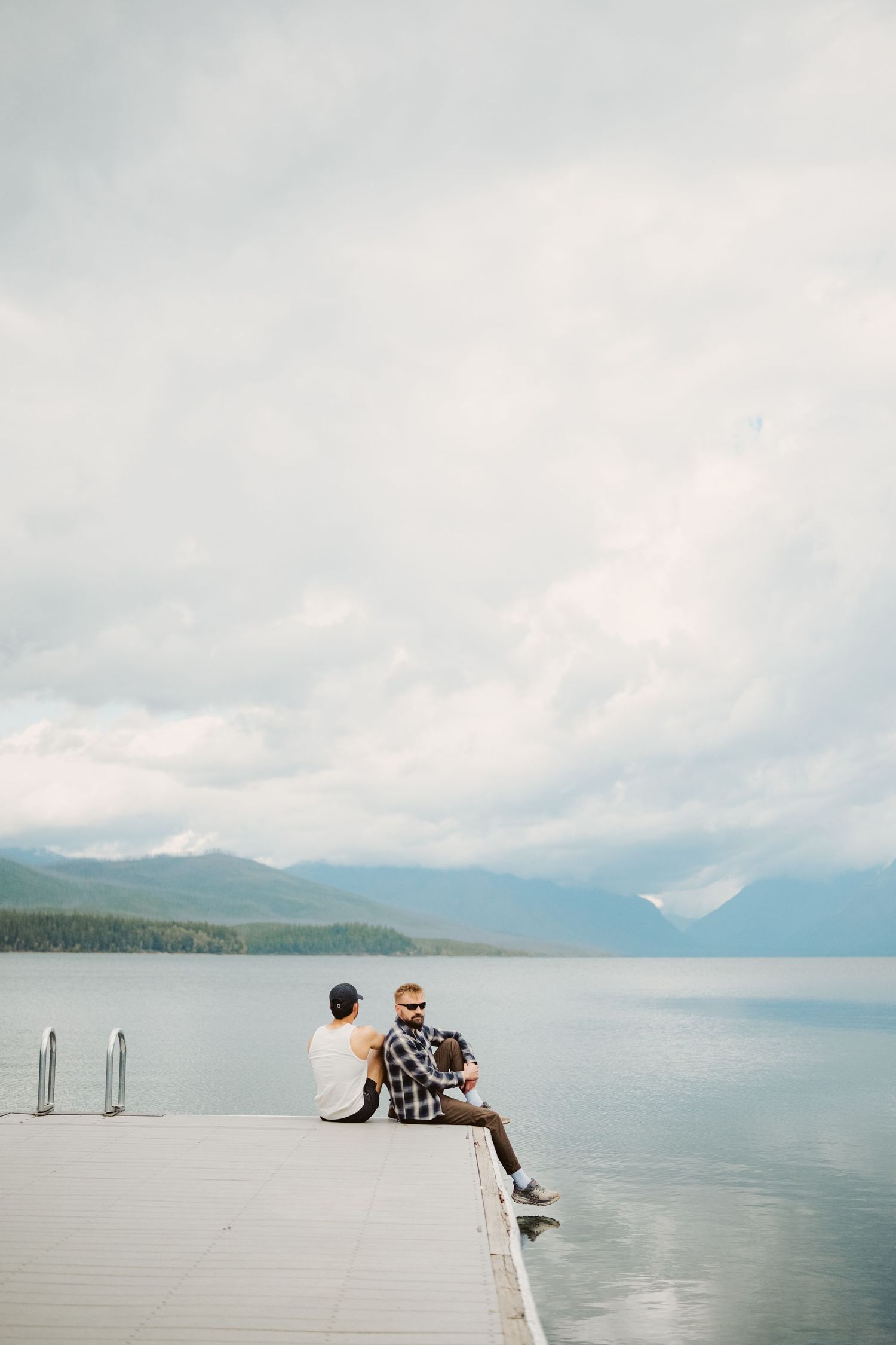 People sit and relax on a wooden dock overlooking a serene mountain lake with sailboats in the distance.