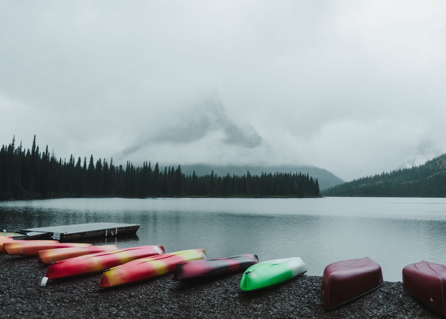 Colorful kayaks line the misty shoreline of a serene mountain lake with evergreen forests in the background.