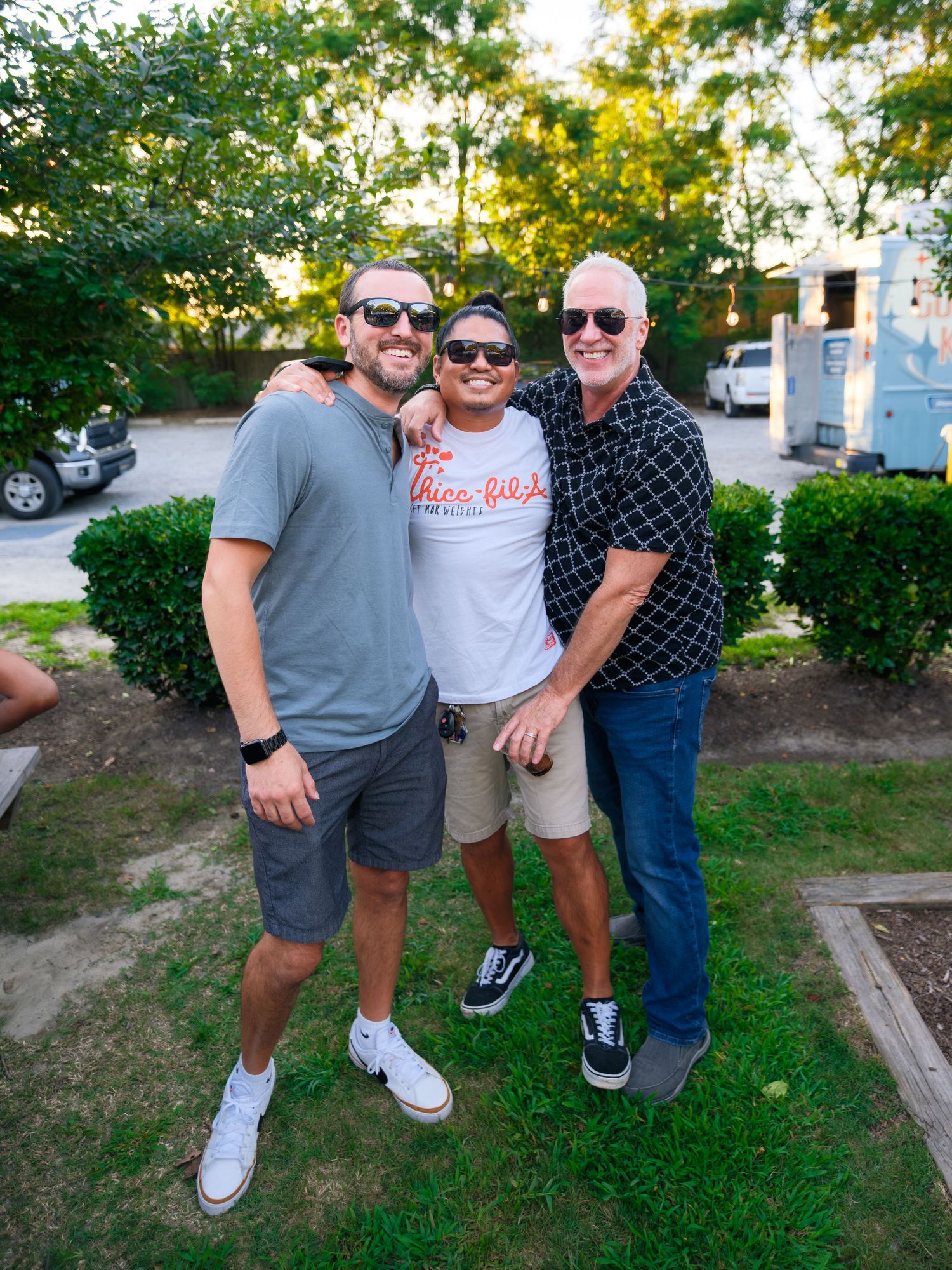 A casual outdoor gathering of three people standing on grass near a building during golden hour.