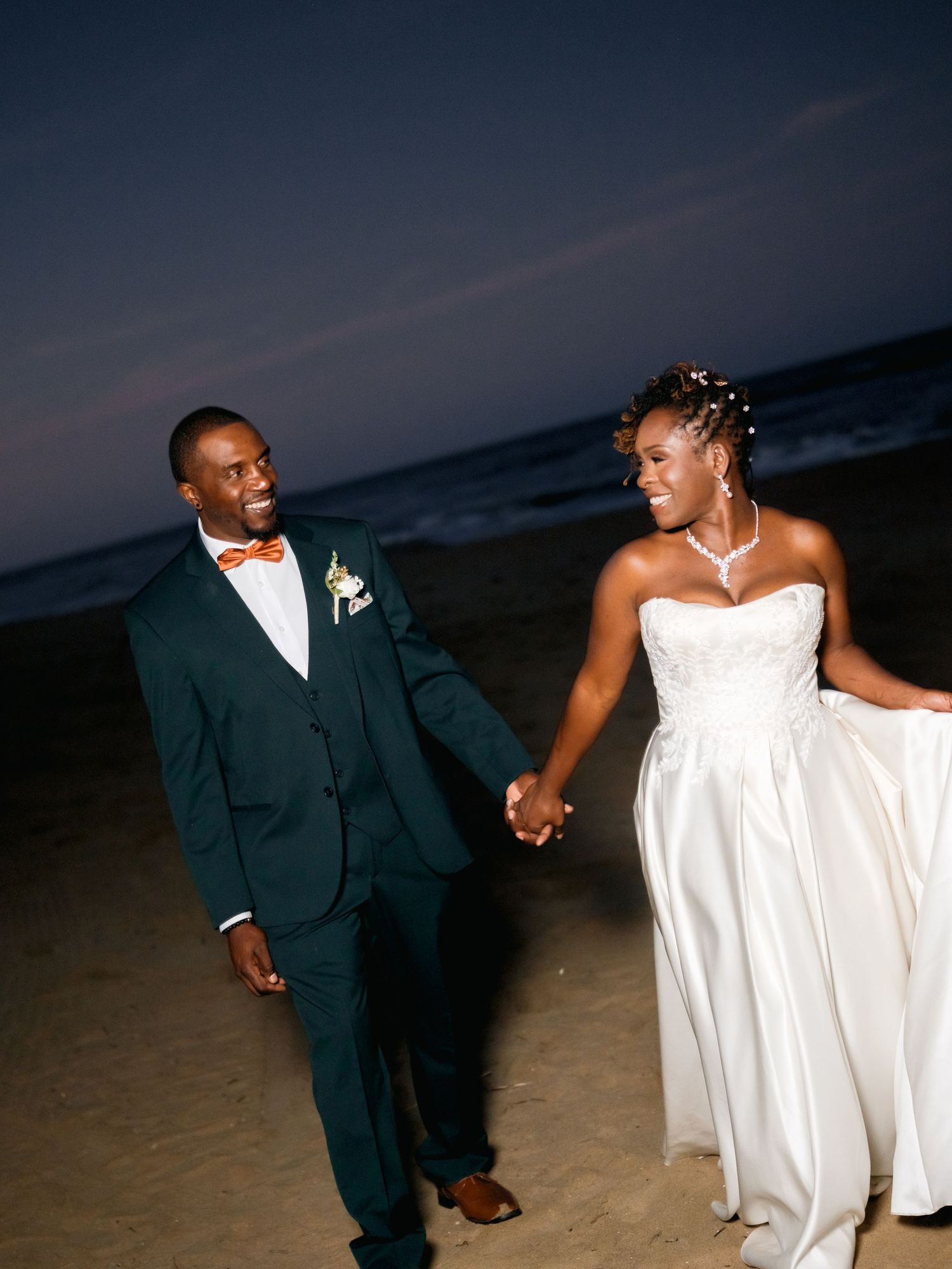 A couple walks hand in hand on the beach at dusk in formal wedding attire.