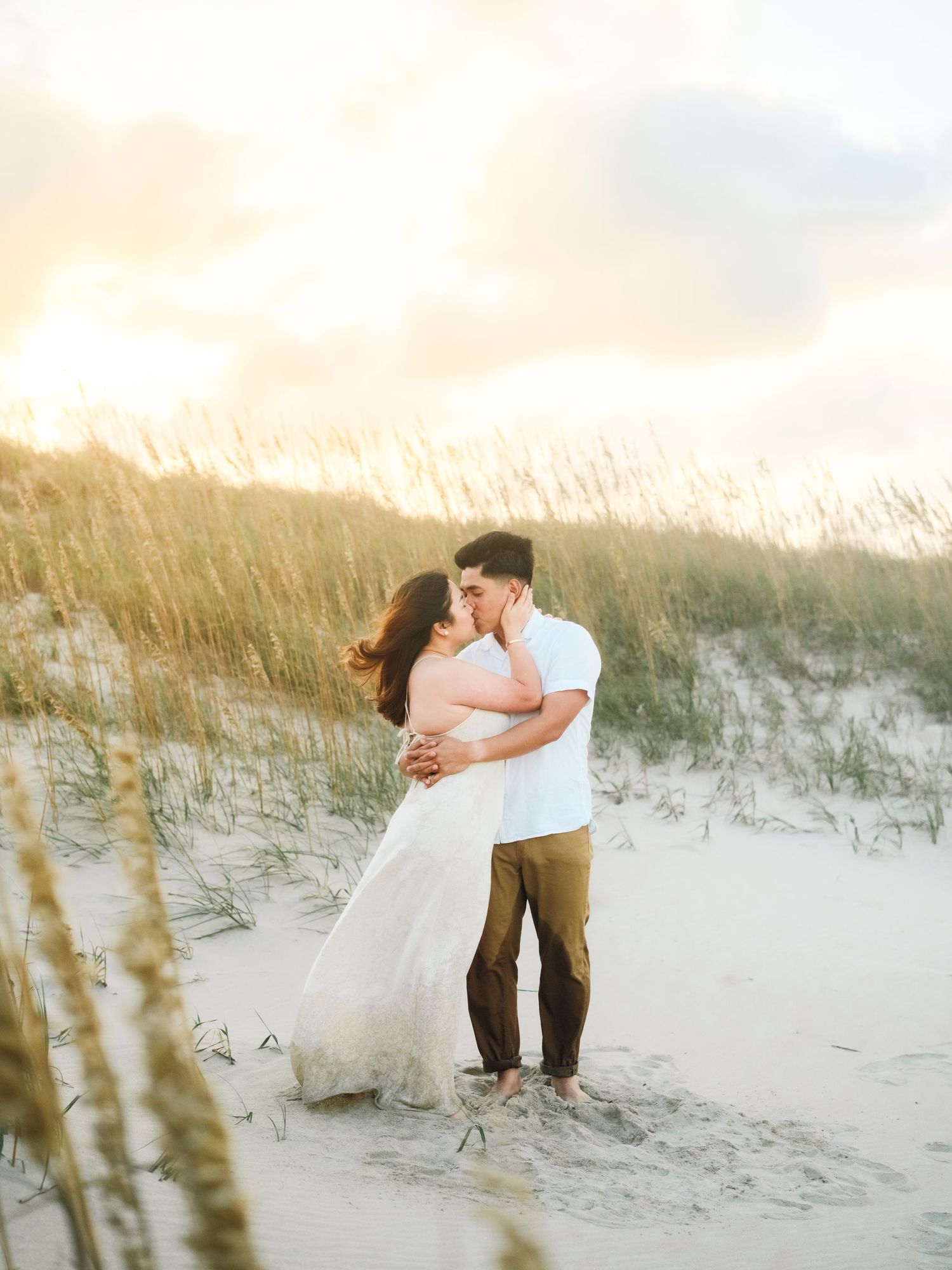 Romantic couple embraces on sandy dunes at sunset with coastal grass swaying in the golden light.