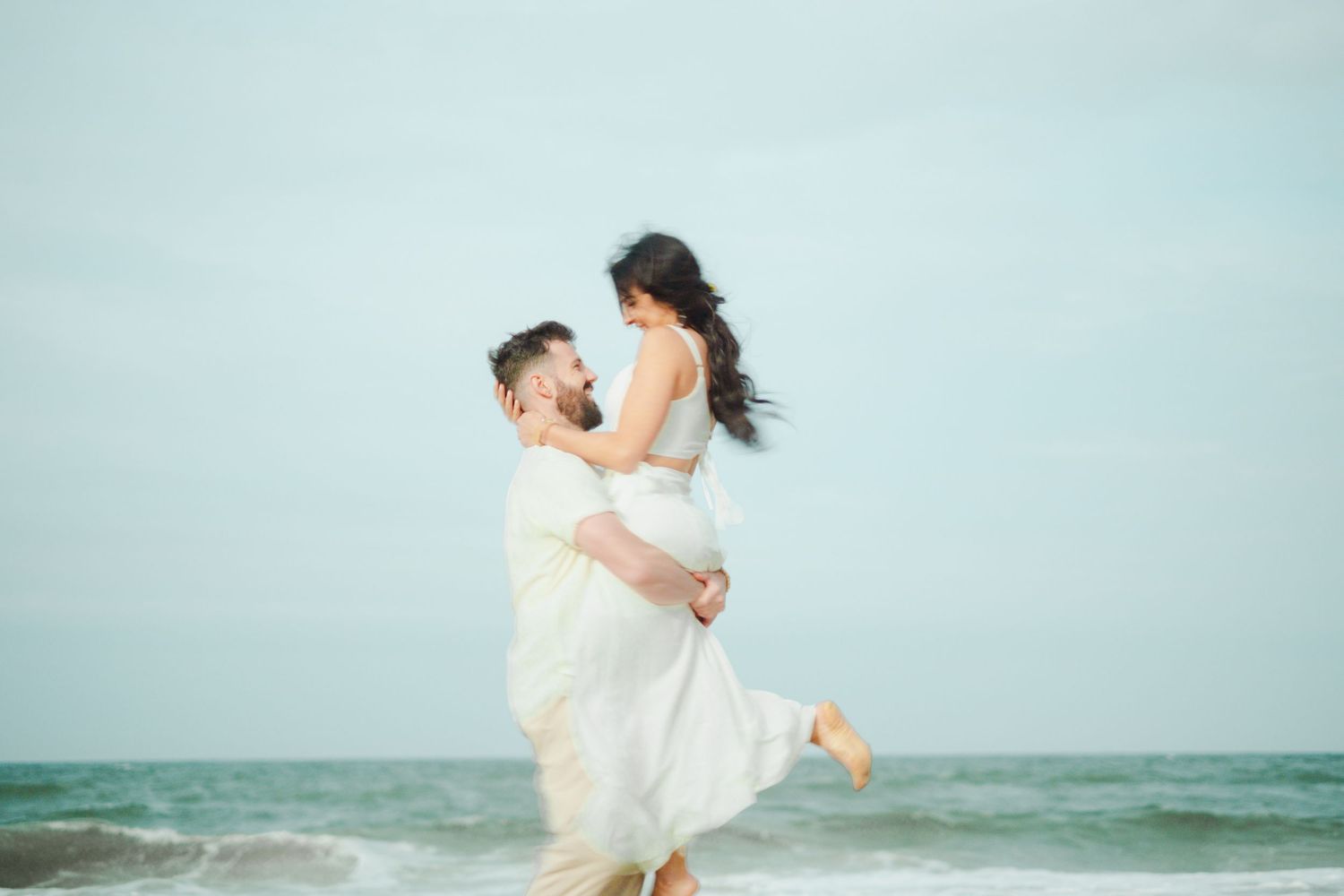 Joyful newlyweds celebrate together against a dreamy oceanfront backdrop, her white dress flowing in the sea breeze.