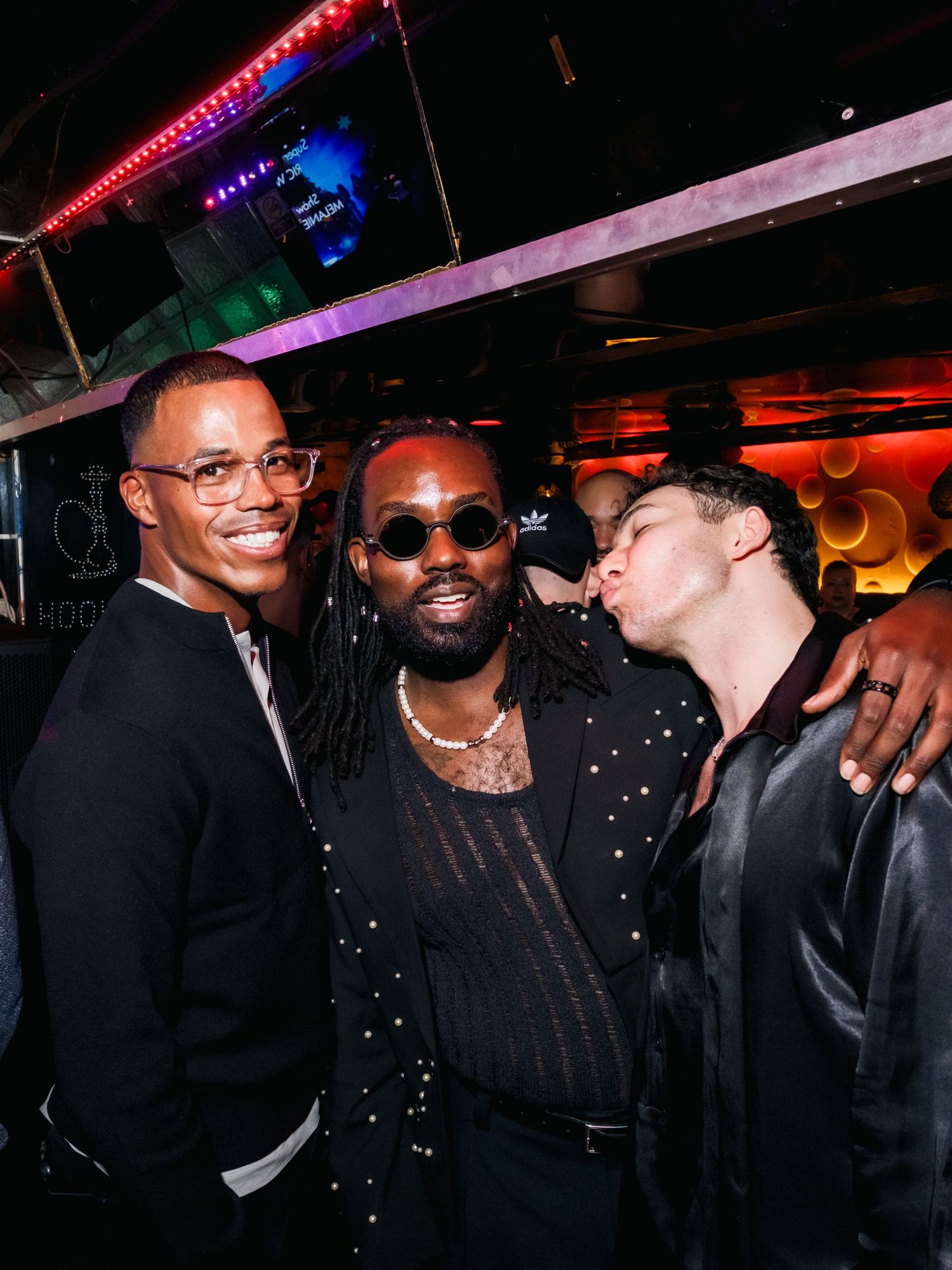 Three friends smiling and posing together at a nightclub with colorful lighting and dark ambiance in the background.