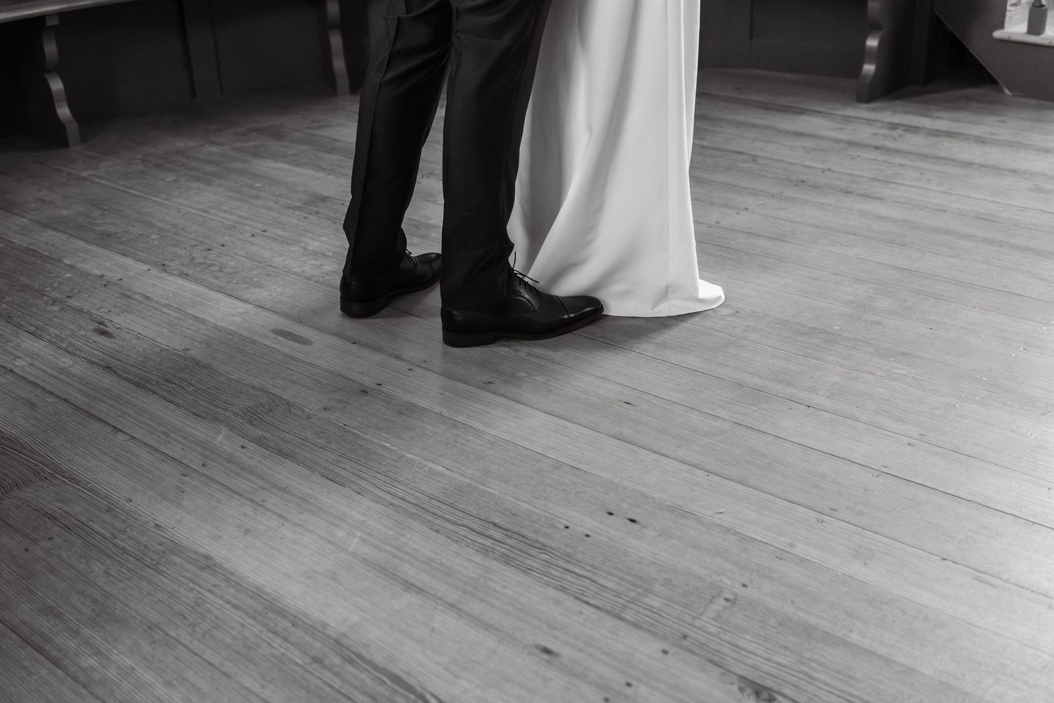 Black and white photo of dress and suit shoes standing close together on a wooden dance floor.