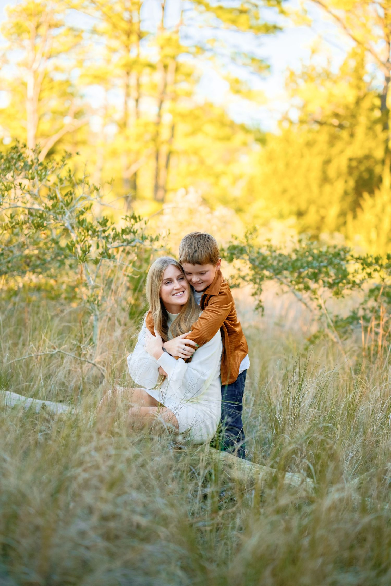 A tender embrace shared between two people in a sunlit meadow with tall grass and golden autumn trees in the background.