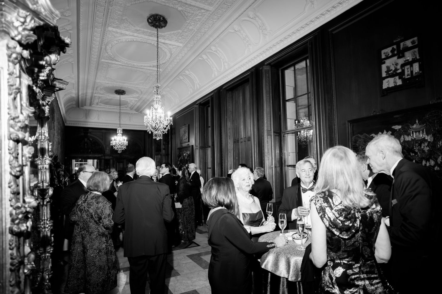 A black and white wedding reception scene with guests mingling under elegant chandeliers in a vintage-style venue.