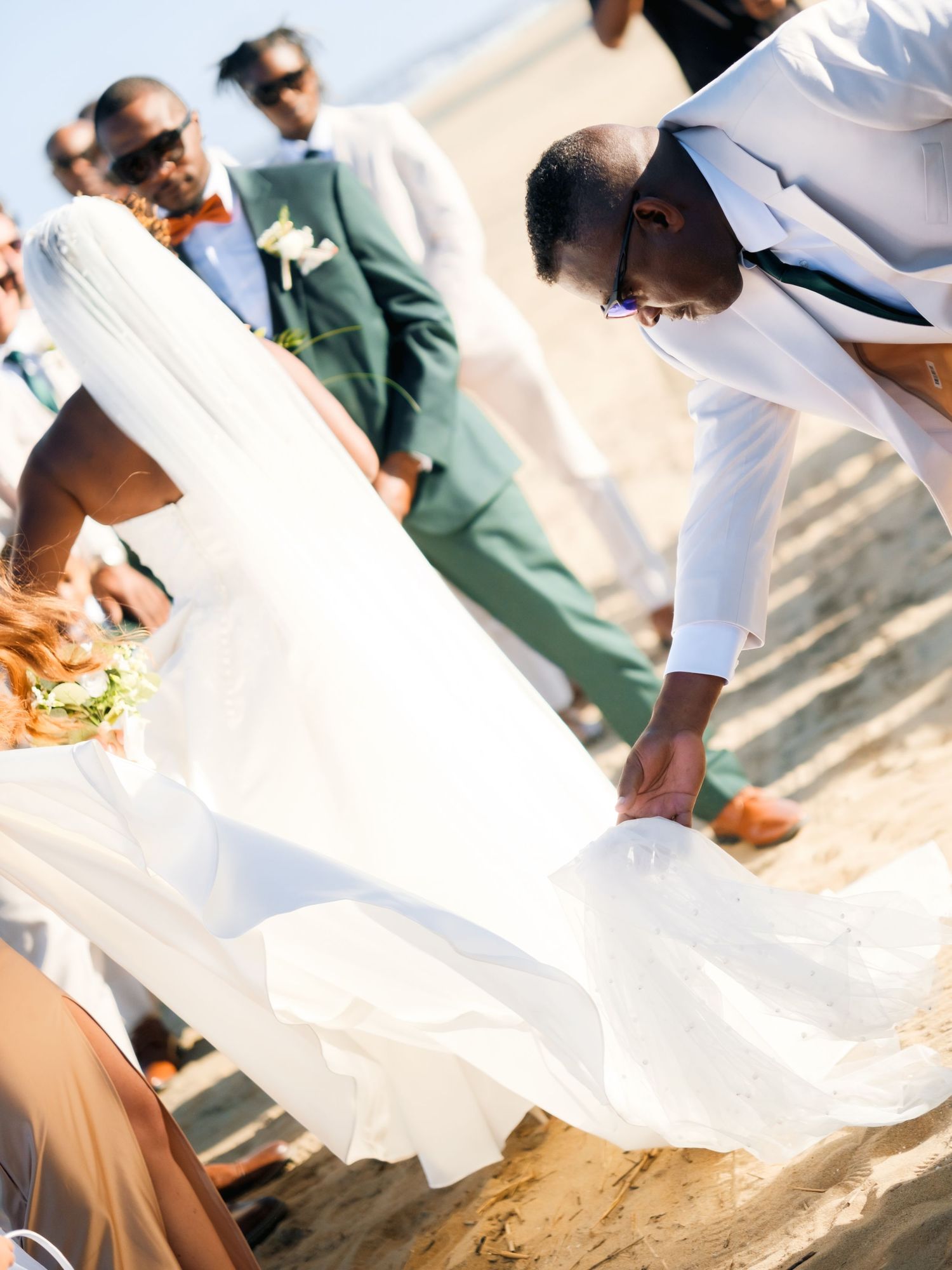 Someone adjusts a white wedding dress train on the beach while others look on.