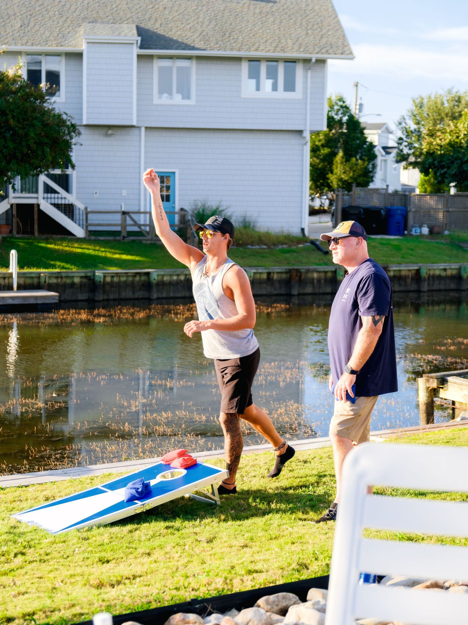 People waving goodbye on a grassy lawn next to a waterfront house on a sunny summer day.