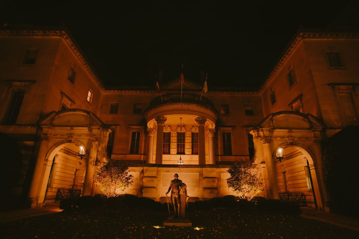 Illuminated historic mansion facade glows warmly at night with ornate architectural details and landscaping visible.