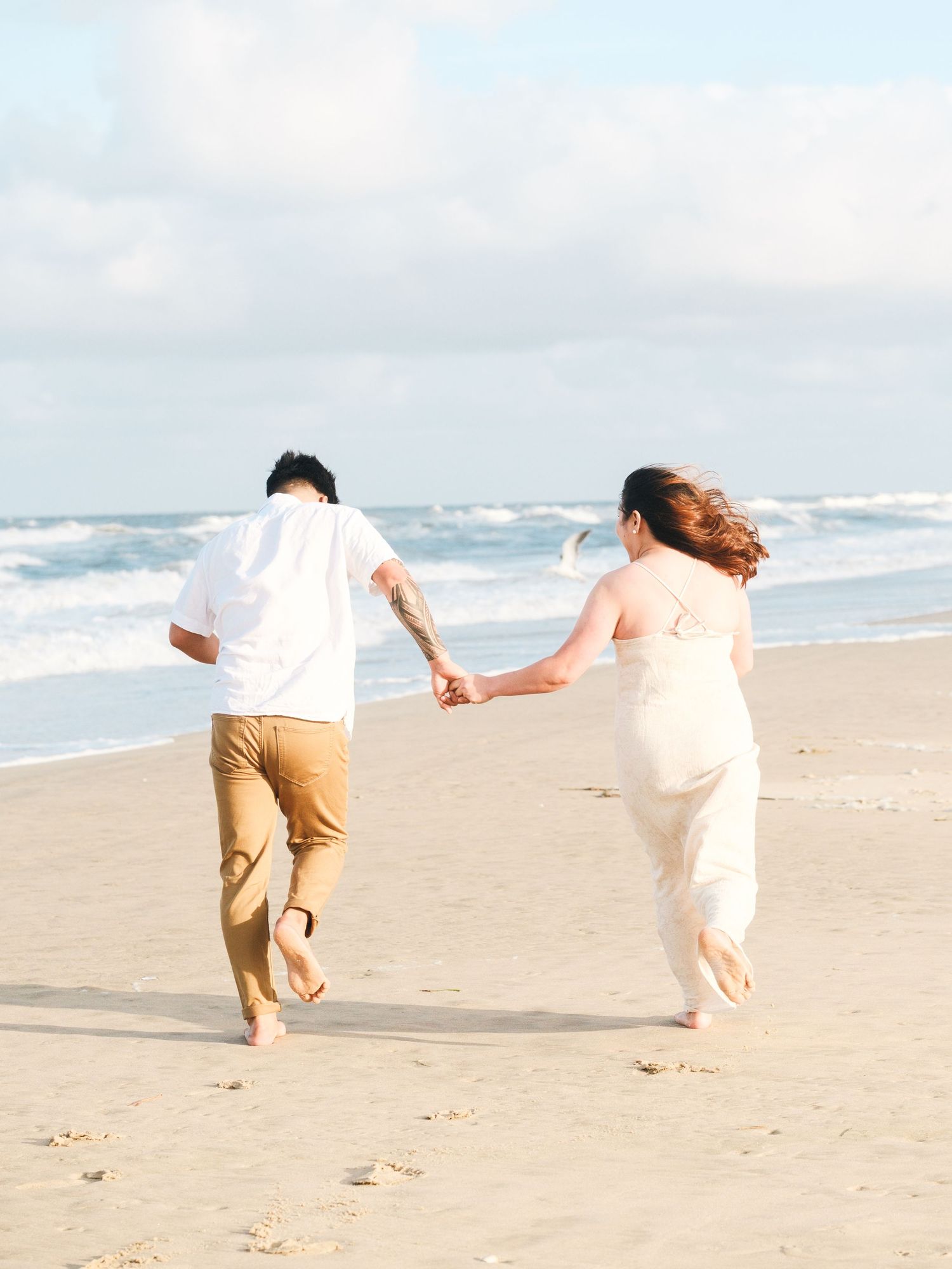 A couple runs playfully along a sandy beach with ocean waves in the background, creating a romantic seaside moment.