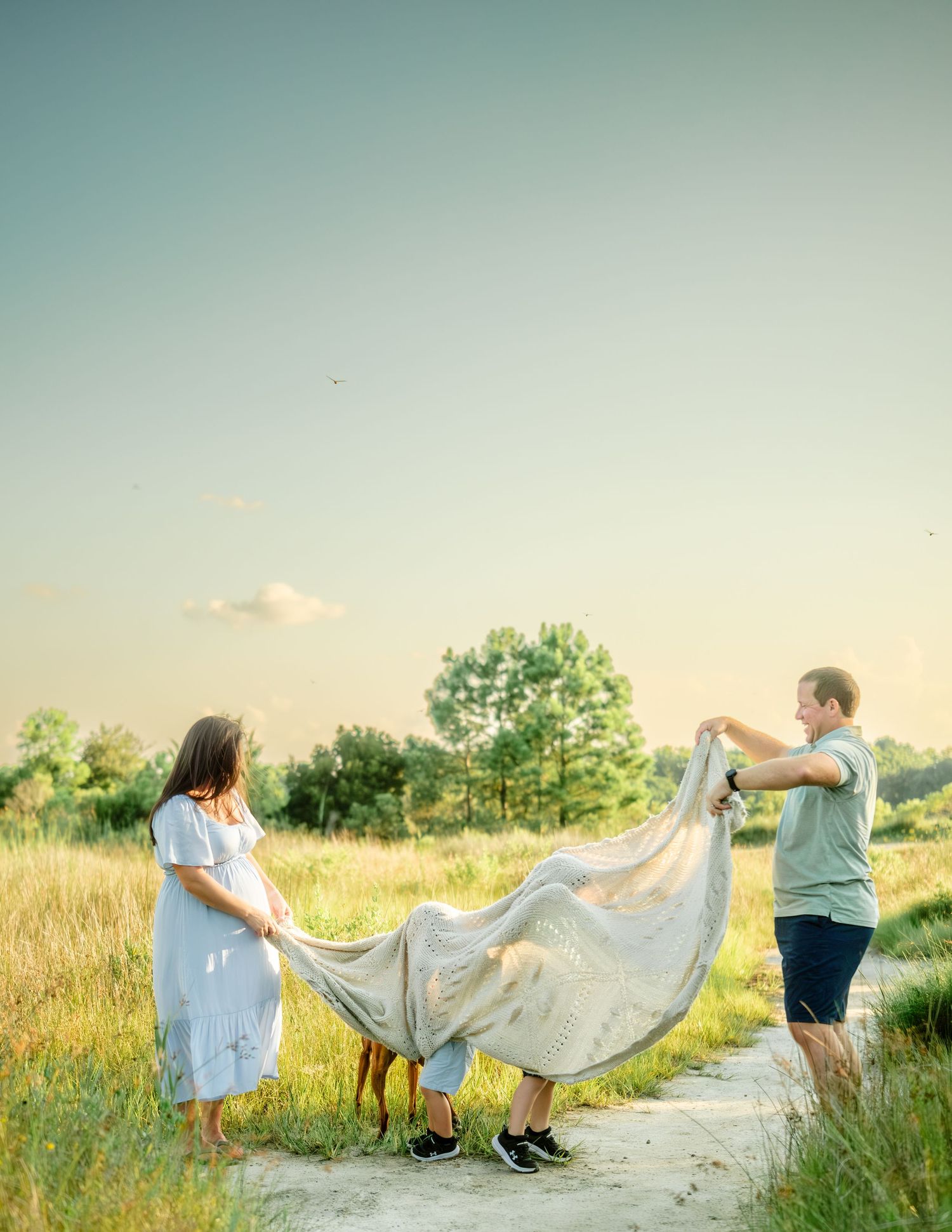 Couple shaking out a blanket together on a dirt path through a summer meadow at sunset.