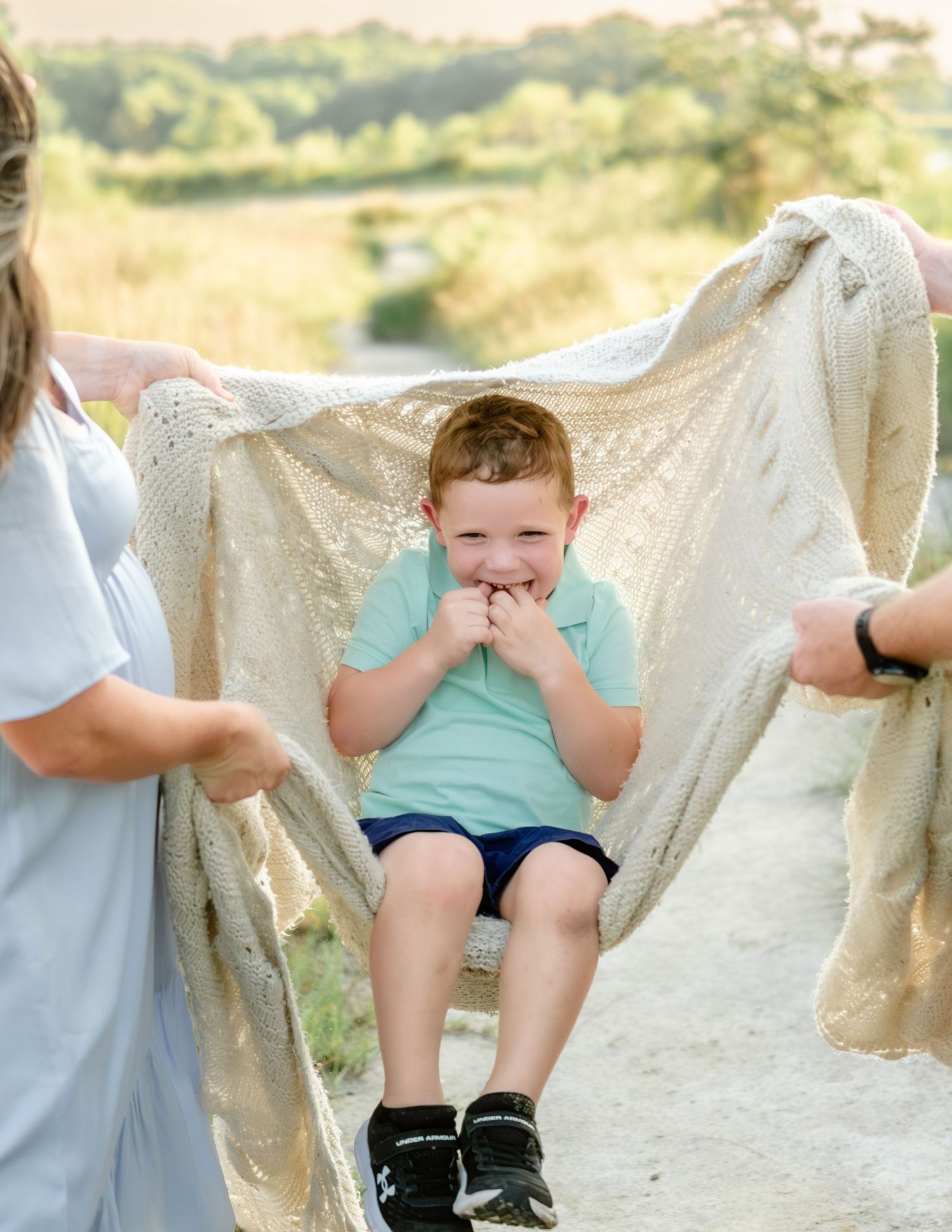 A child in a turquoise shirt laughs while seated between two people holding up a flowing cream-colored fabric outside.