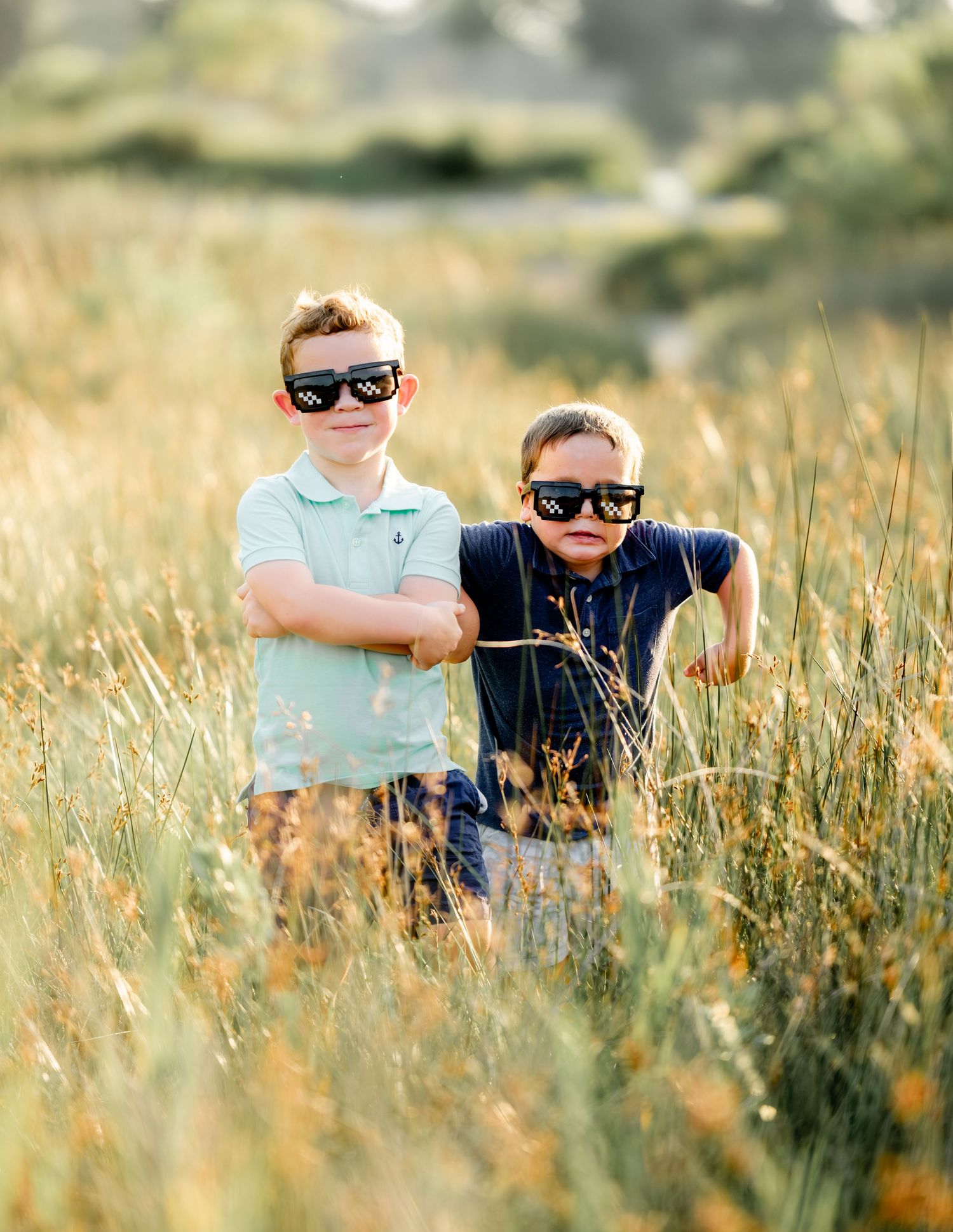 Two children wearing sunglasses stand together in a tall golden field of grass on a sunny summer day.