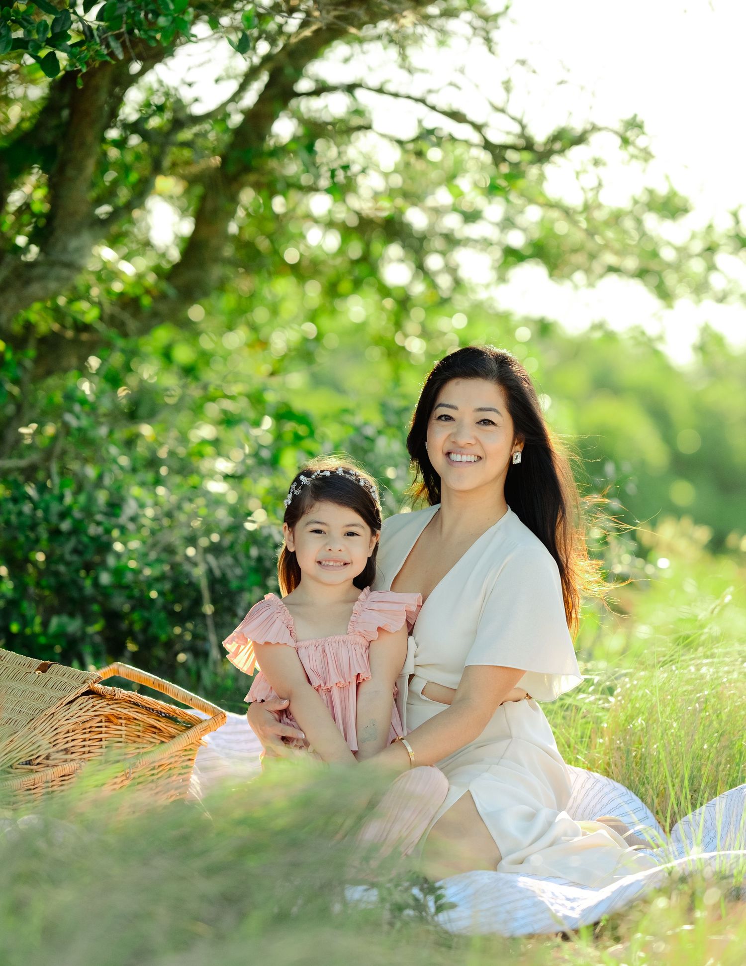 Two people enjoying a sunny outdoor picnic with a basket on a blanket surrounded by lush green trees and grass.