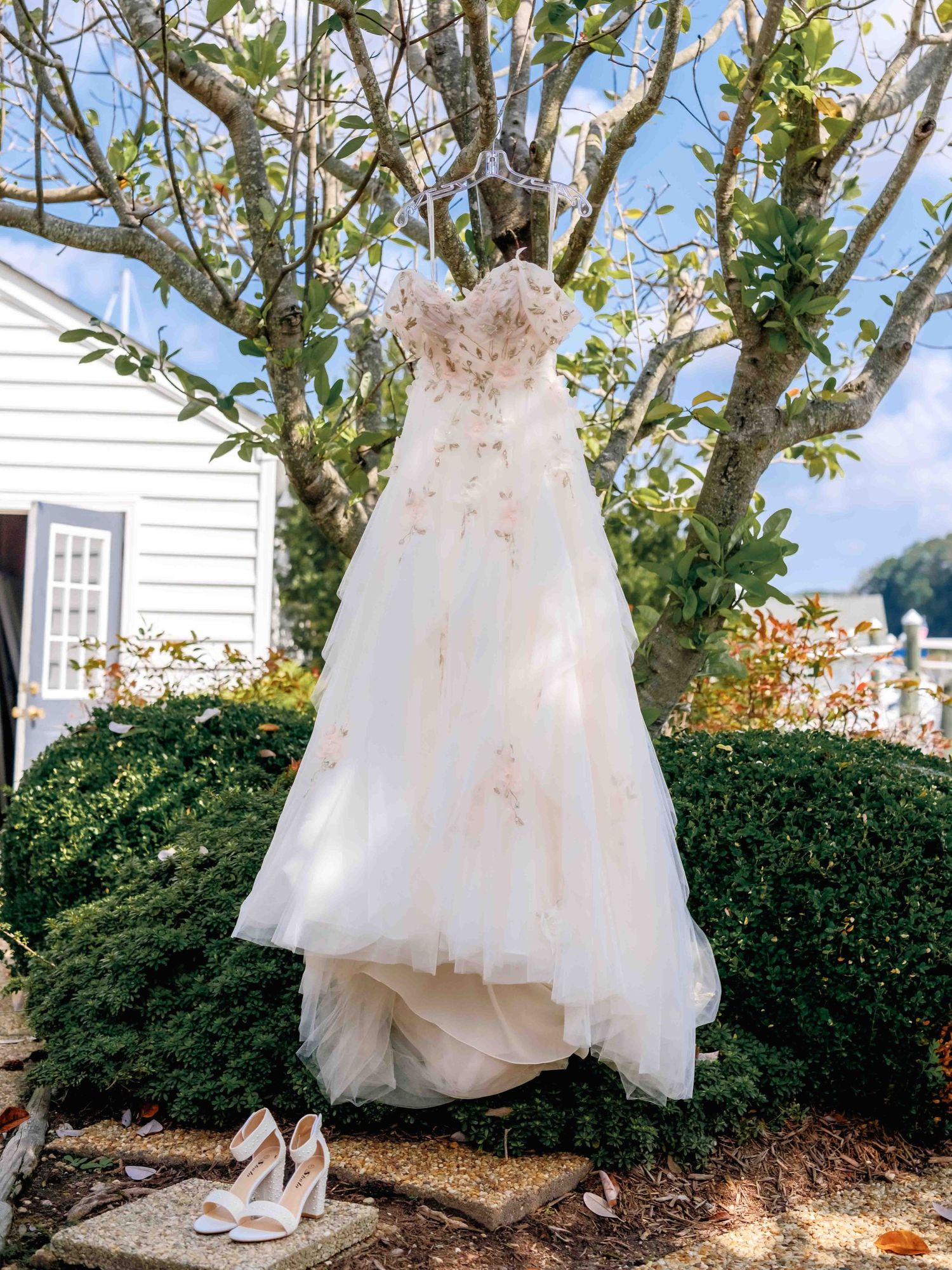 A delicate white wedding dress and matching heels displayed outdoors against greenery and a white building exterior.
