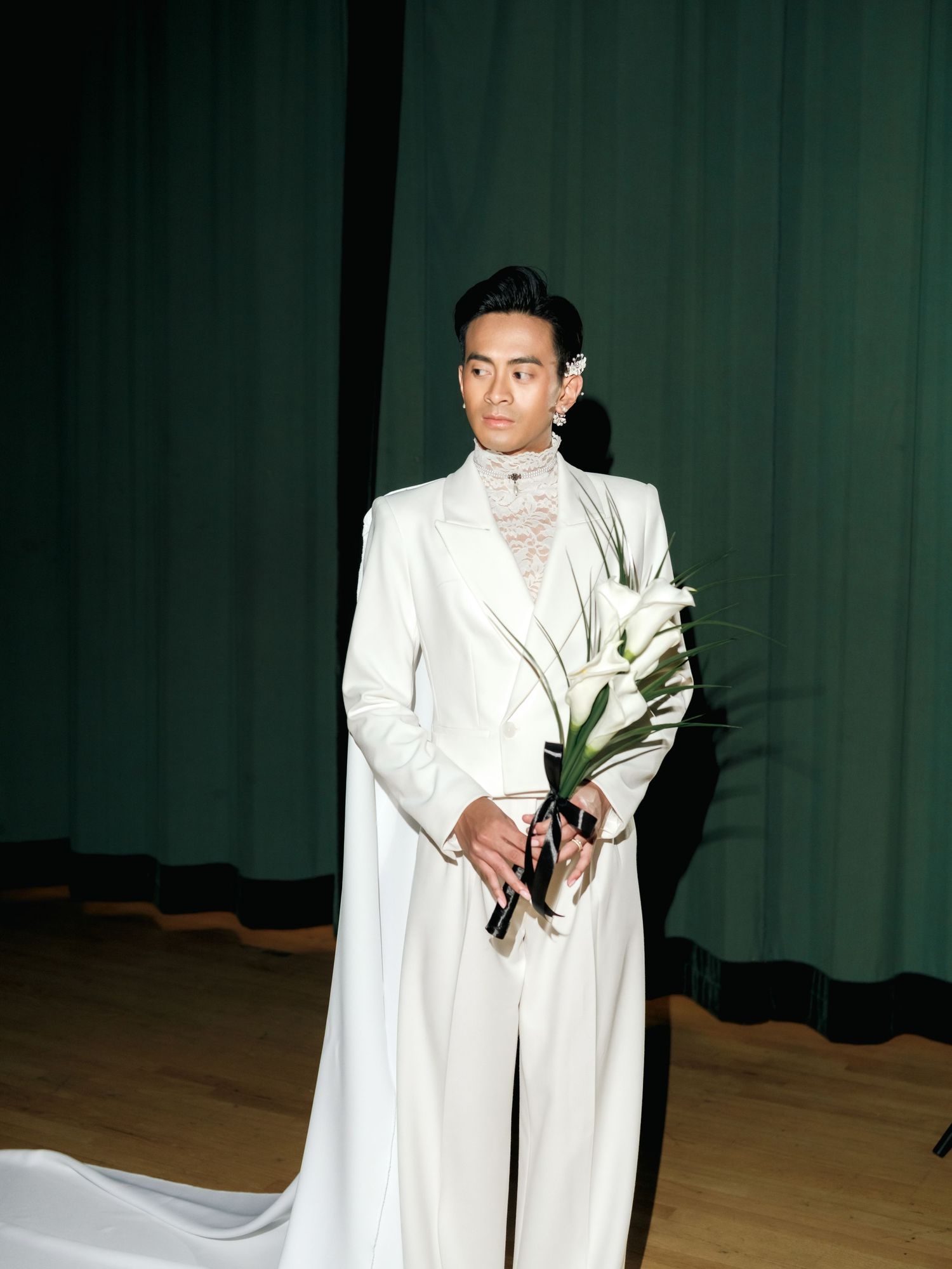 Someone poses in an elegant white formal suit with long train while holding calla lilies against a dark green curtain backdrop.