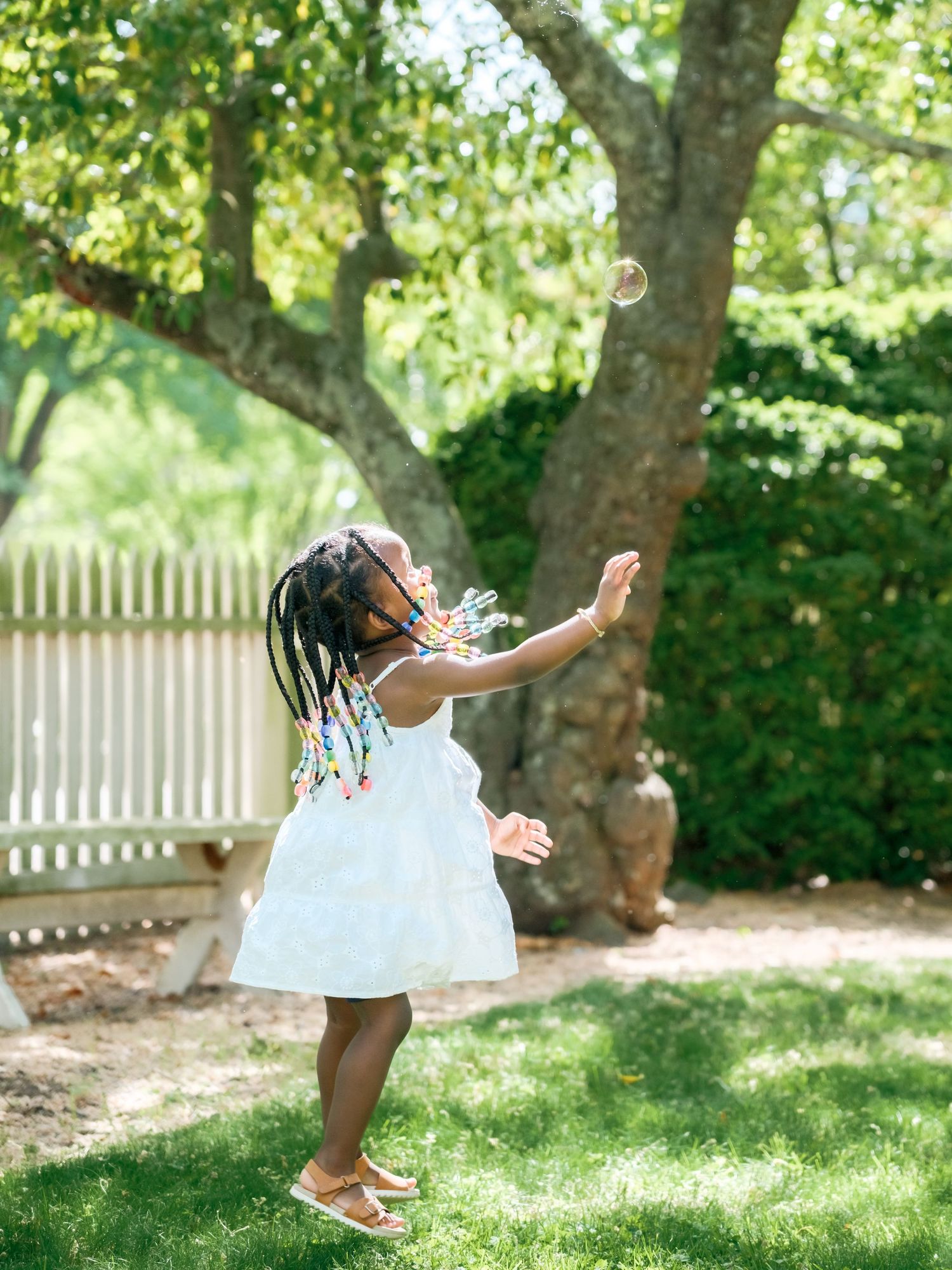 A child in a white dress jumps and plays outside on a sunny day in a backyard with trees and a white fence.