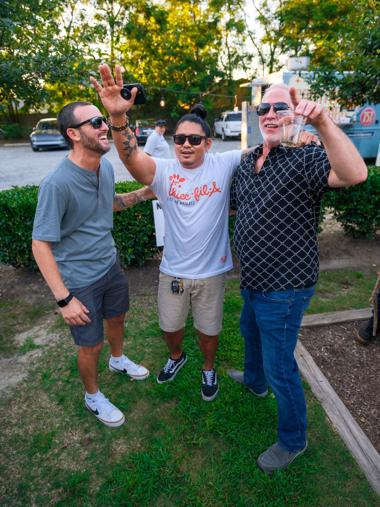 Three people in casual attire stand together outdoors on grass, waving and smiling on a sunny day near parked cars.