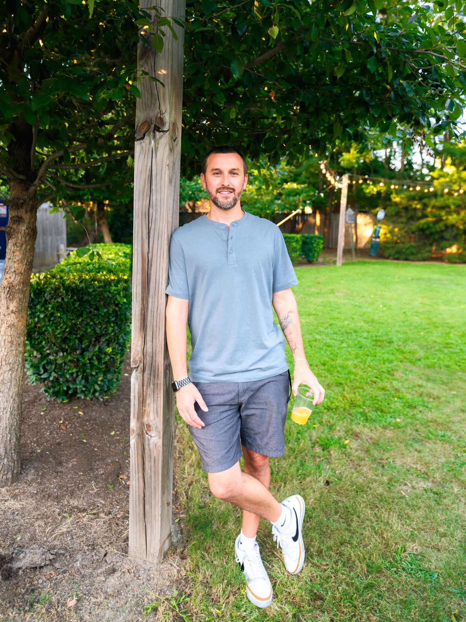 Person wearing a blue t-shirt and gray shorts poses playfully in front of trees in an outdoor garden setting.