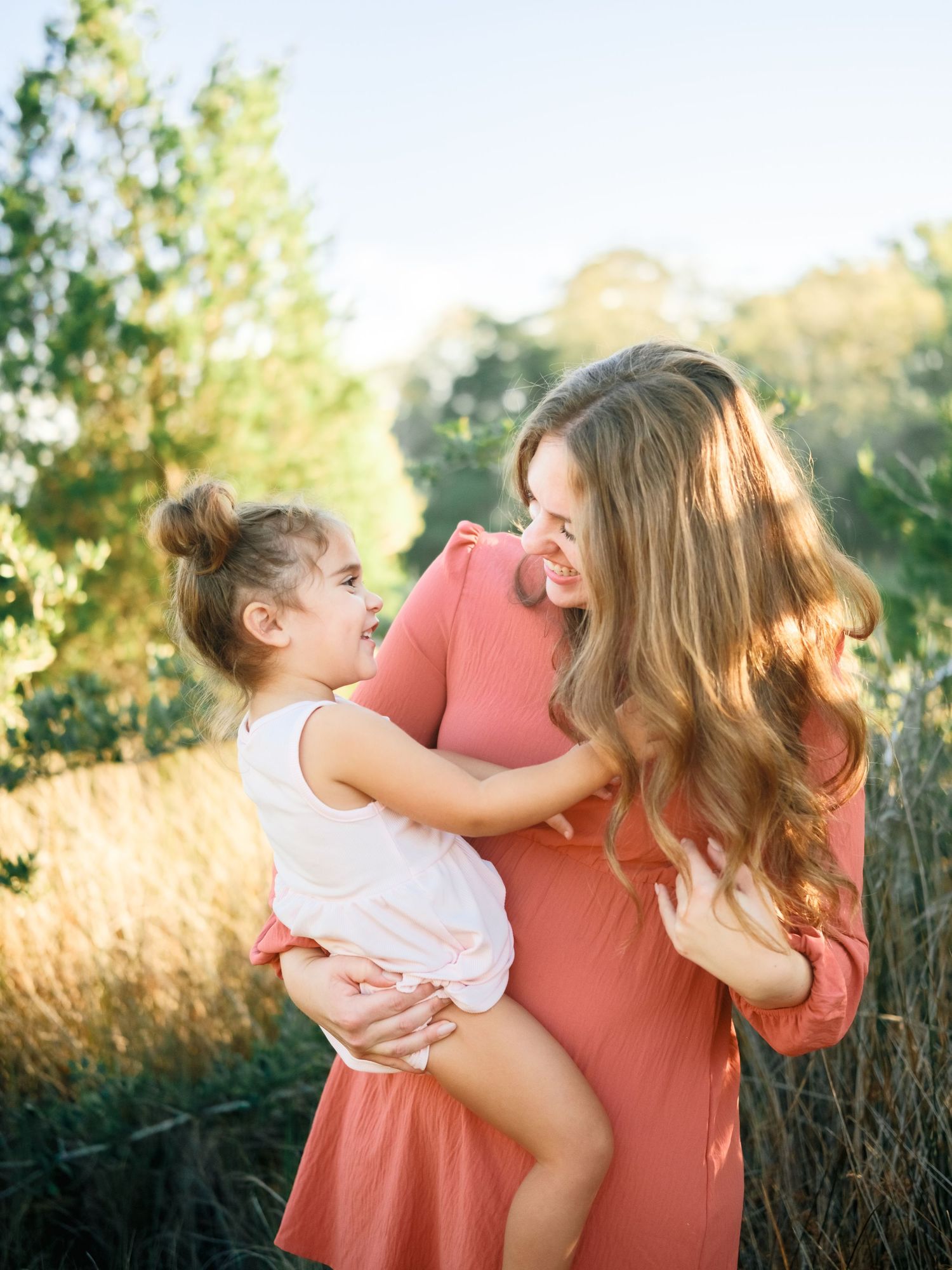 A mother and daughter share joyful moments together in a golden field during sunset, wearing coral and white outfits.