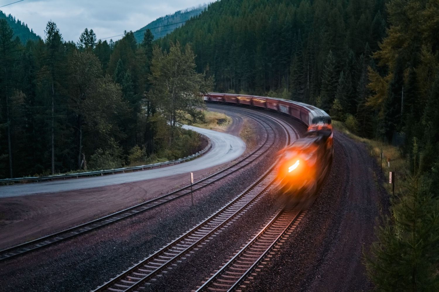 A series of scenic railroad track curves through a forested mountain landscape at dusk with a train's headlight visible.