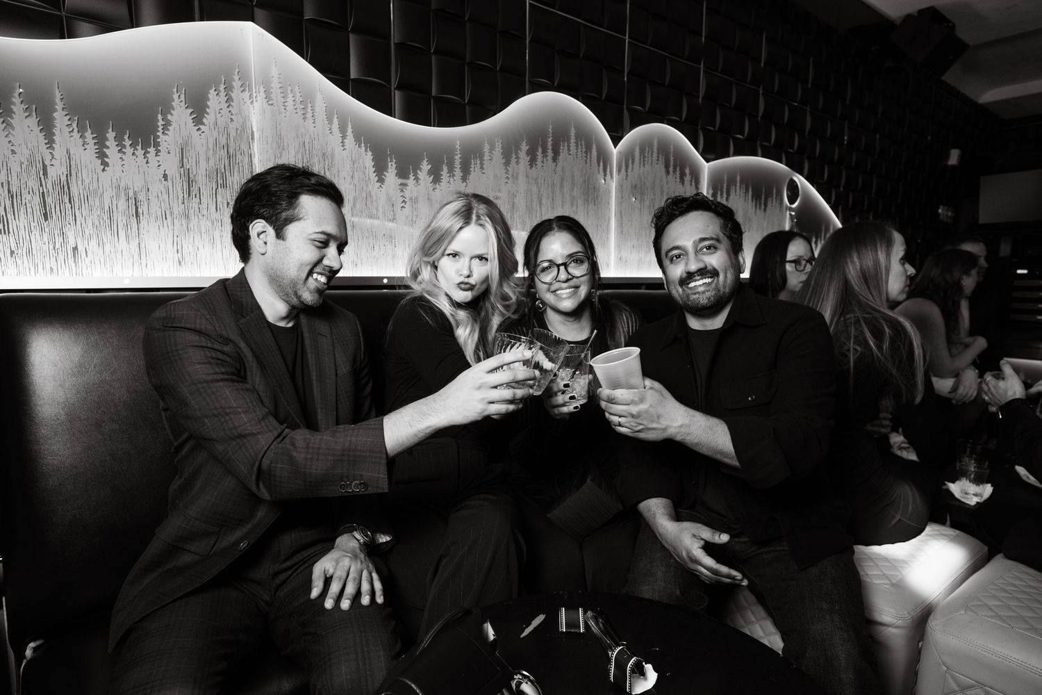 A black and white photo of friends laughing and toasting drinks together at a bar with illuminated wall decor.