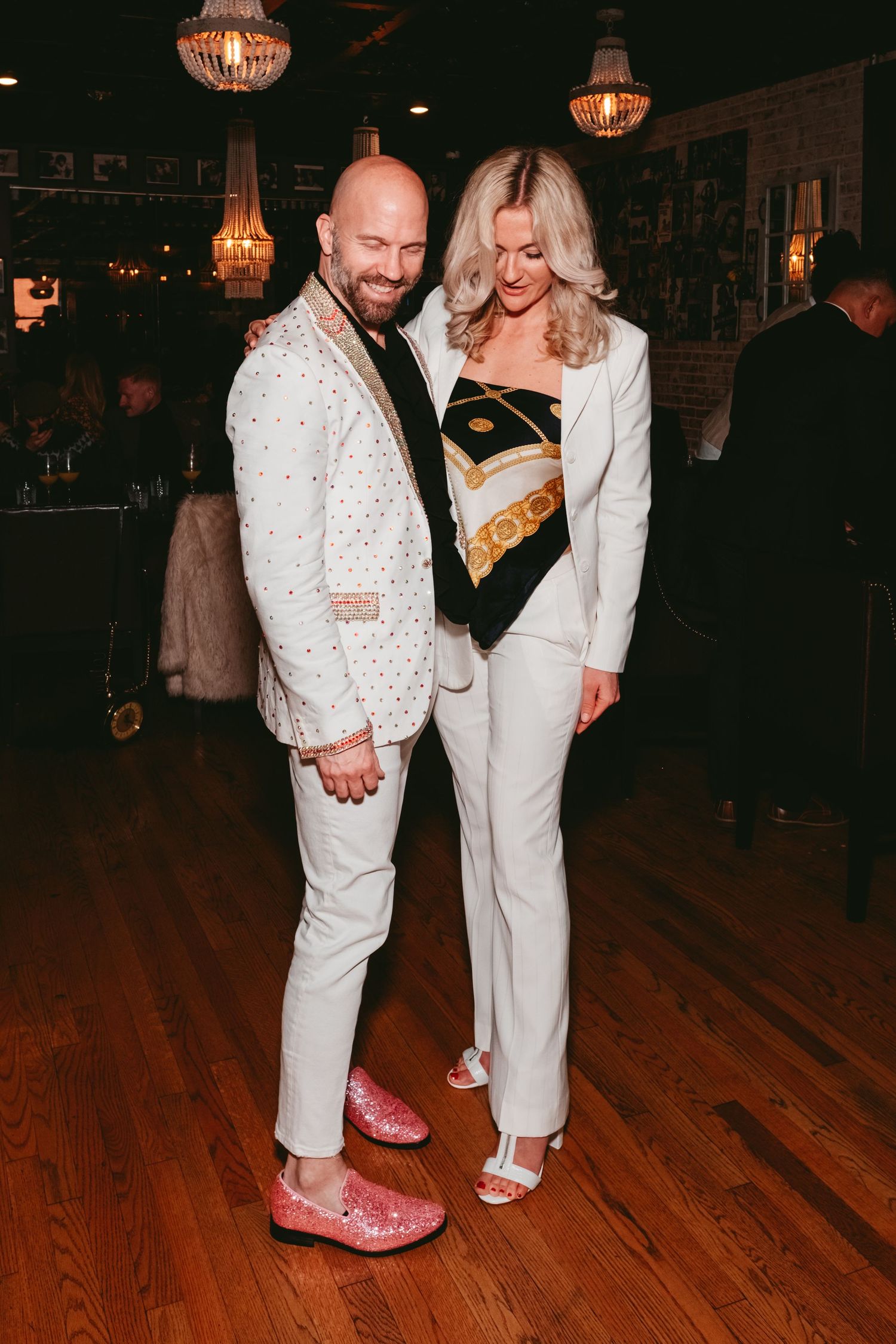 Two people pose together in white outfits on a wooden dance floor under crystal chandeliers at an evening event.