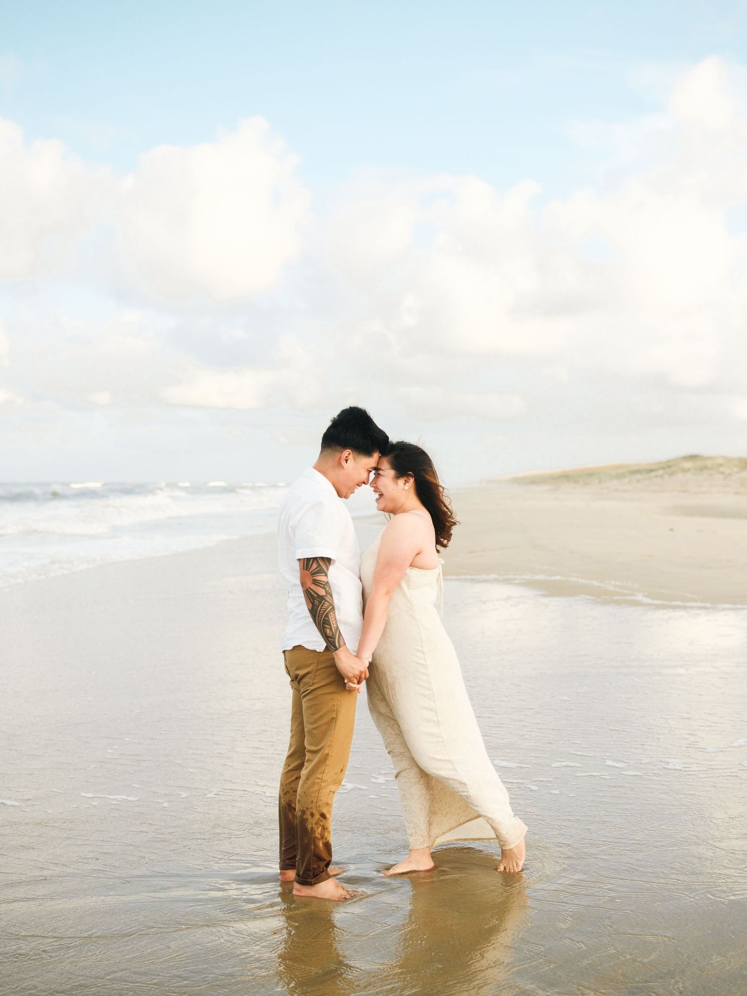 Couple in wedding attire playing in shallow ocean water at sunset on a sandy beach.
