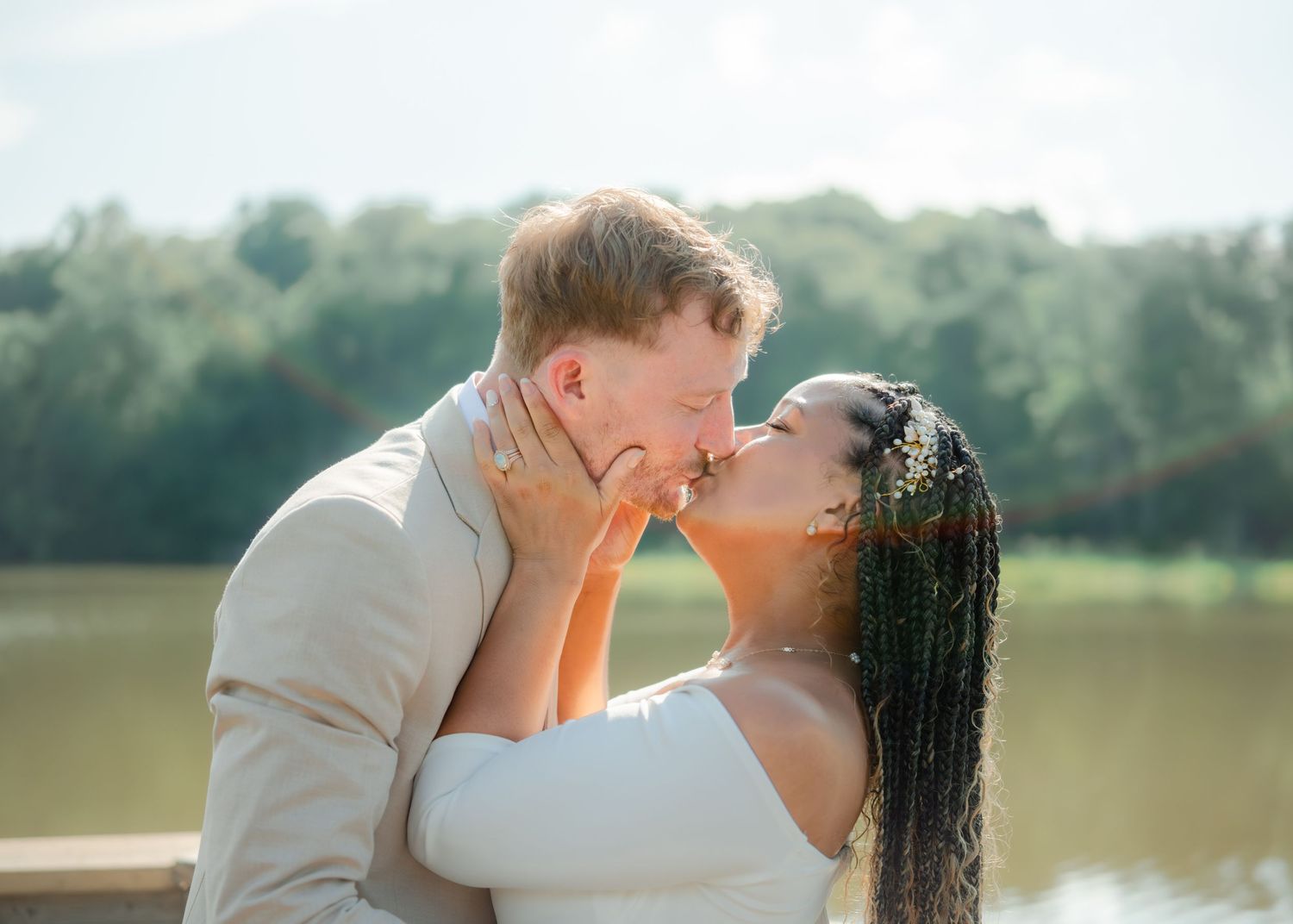 A romantic couple shares a tender kiss by a serene lake with sunlit trees in the background.