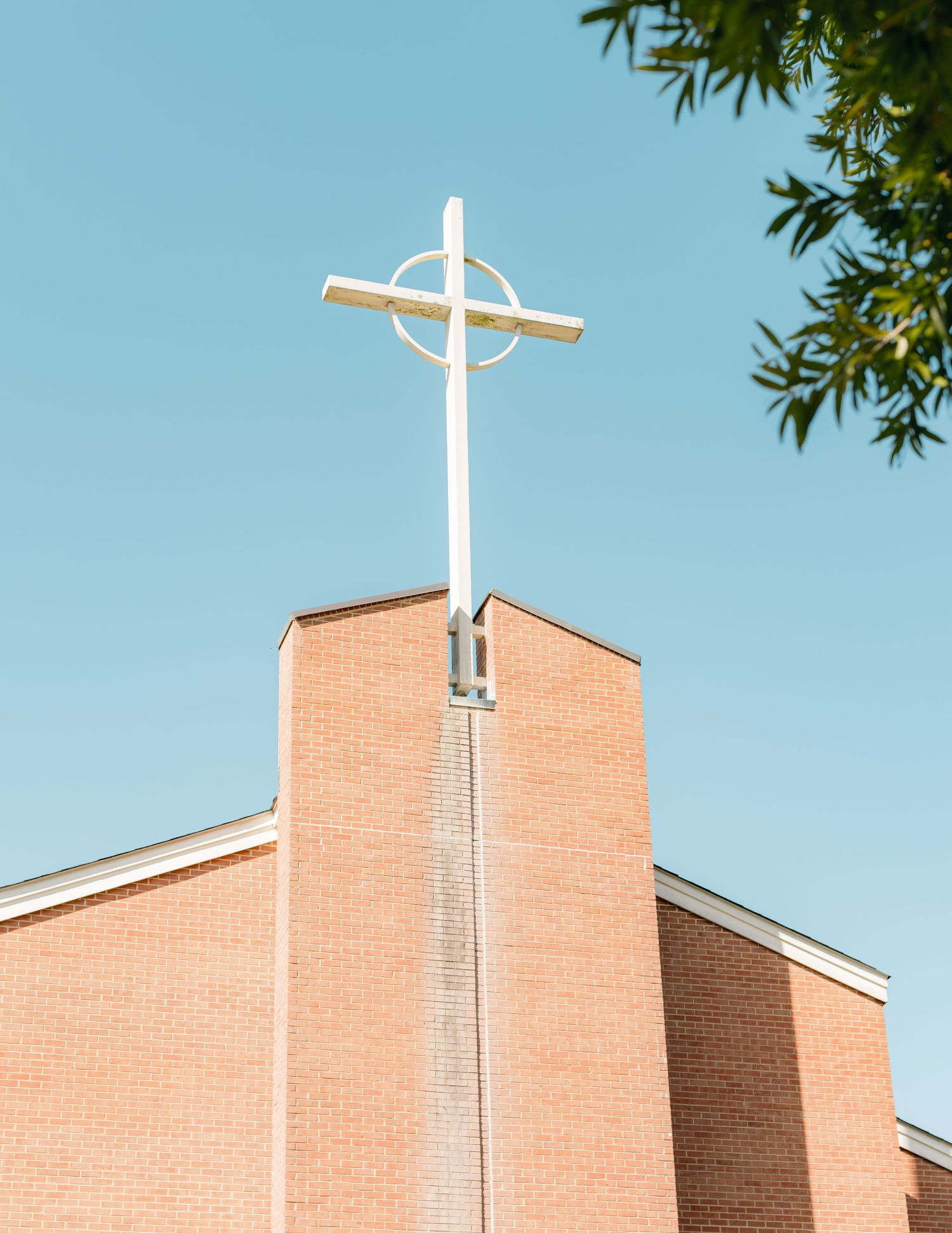 A white cross mounted on top of a modern brick church steeple against a bright blue sky with green leaves in view.