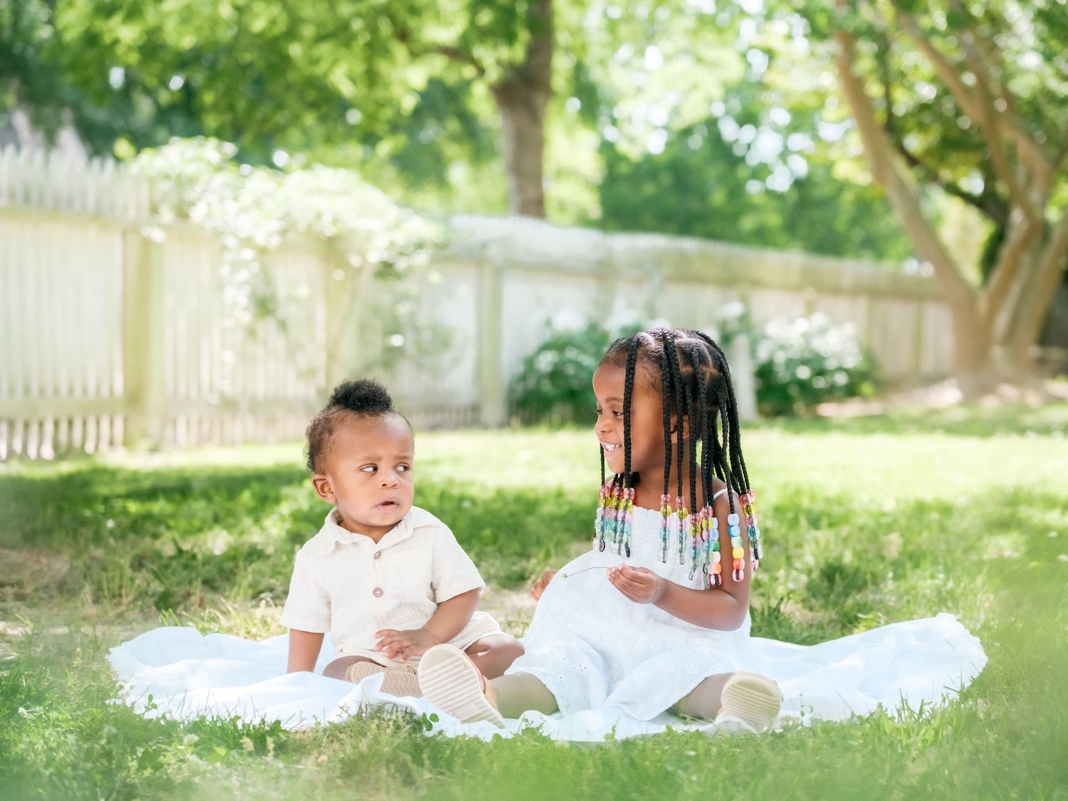 Two children in white outfits sit together on a blanket in a sunny backyard with a white picket fence.